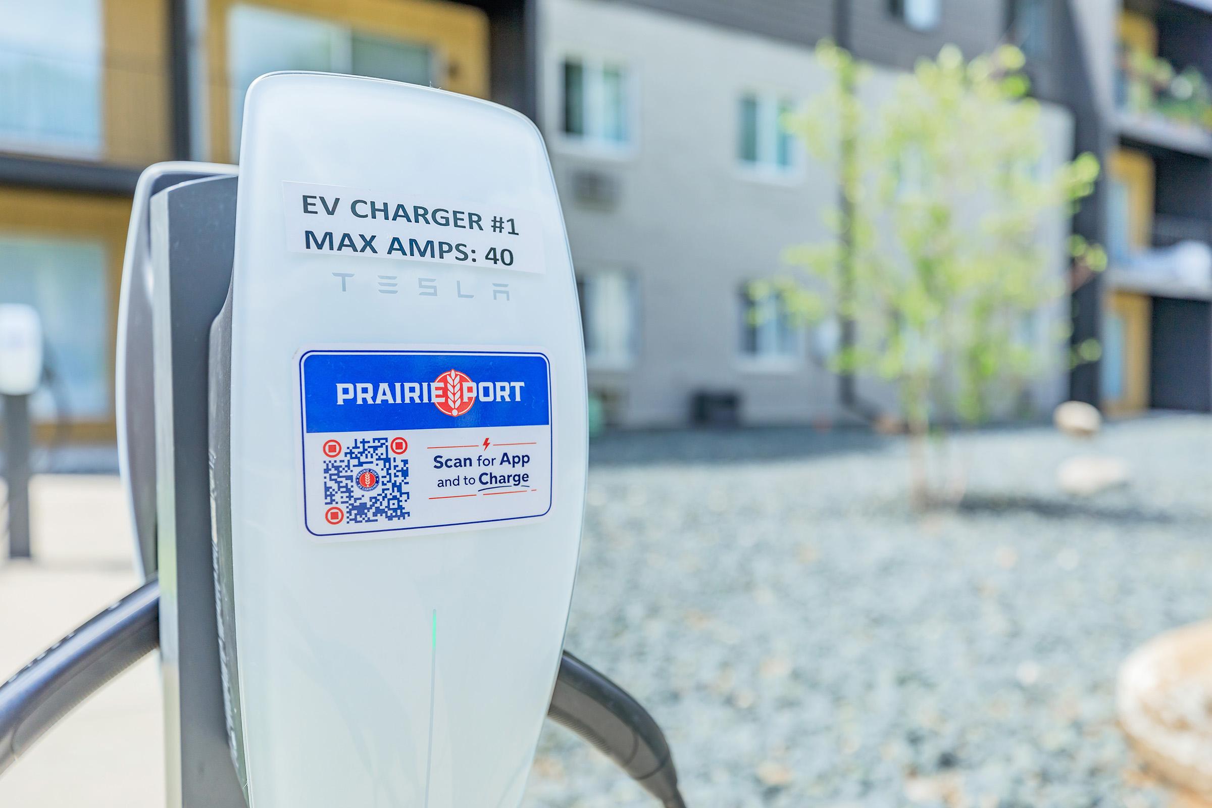 Electric vehicle (EV) charger labeled "Prairie Port" with a QR code for app access. The charger indicates a maximum capacity of 40 amps. In the background, an apartment building is visible, surrounded by gravel and a few sparse plants.