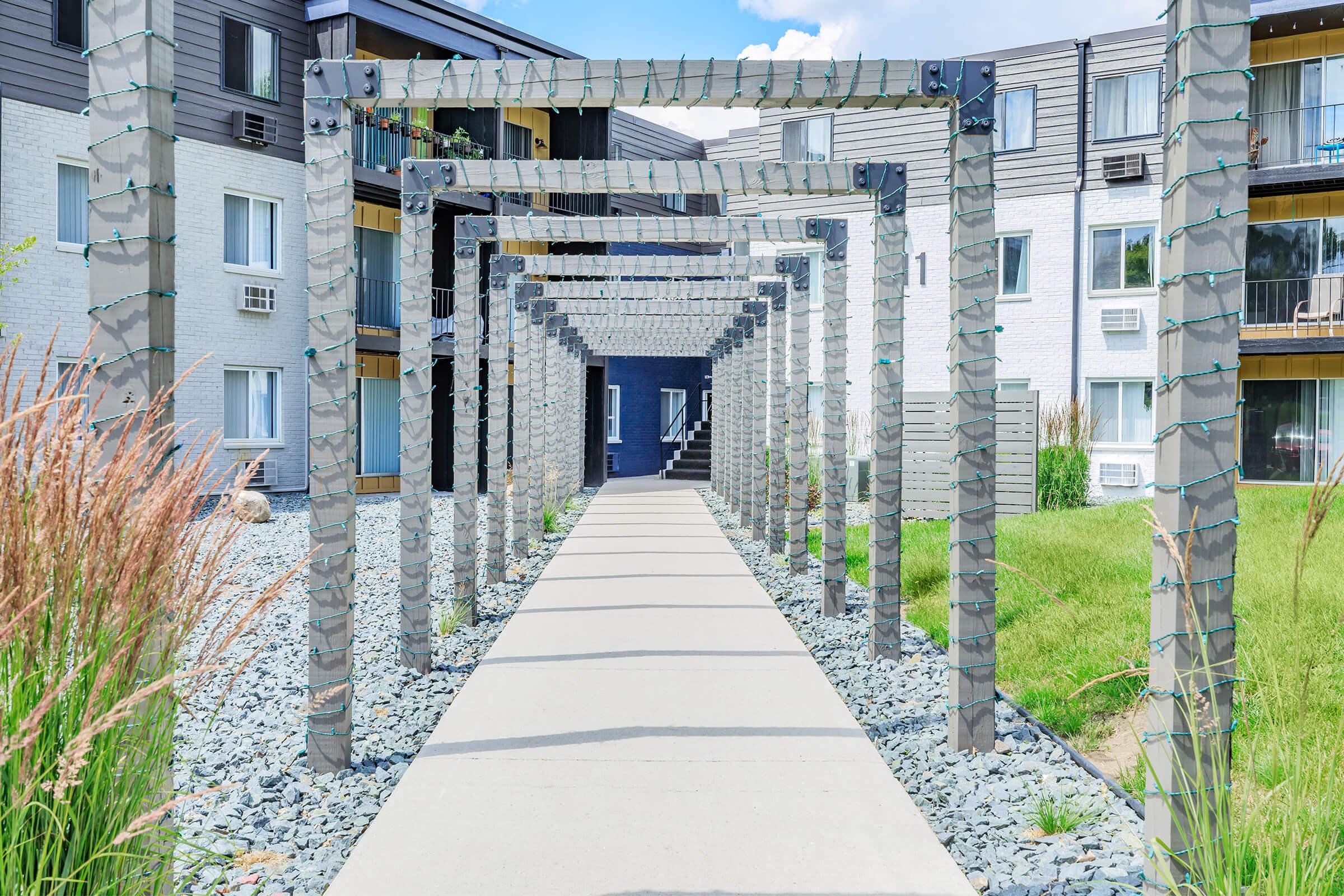 A pathway lined with decorative lighted arches leads through a landscaped area with grass and gravel. In the background, modern apartment buildings are visible, showcasing a mix of textures and colors under a clear blue sky. The scene is bright and inviting, creating a welcoming atmosphere.