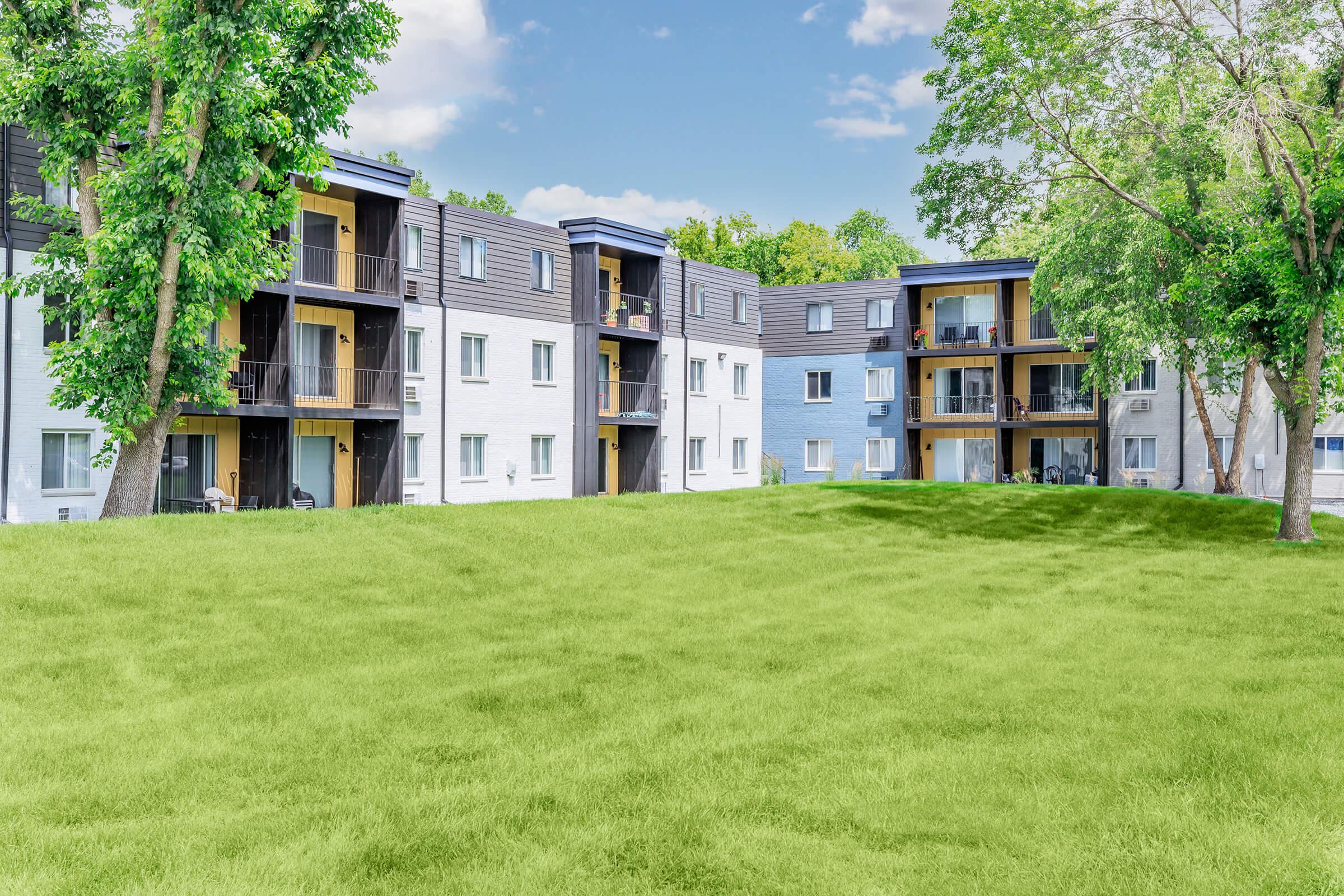 A modern apartment complex with three-story buildings featuring balconies, surrounded by lush green grass and trees under a blue sky with fluffy clouds. The scene conveys a serene and inviting residential atmosphere.