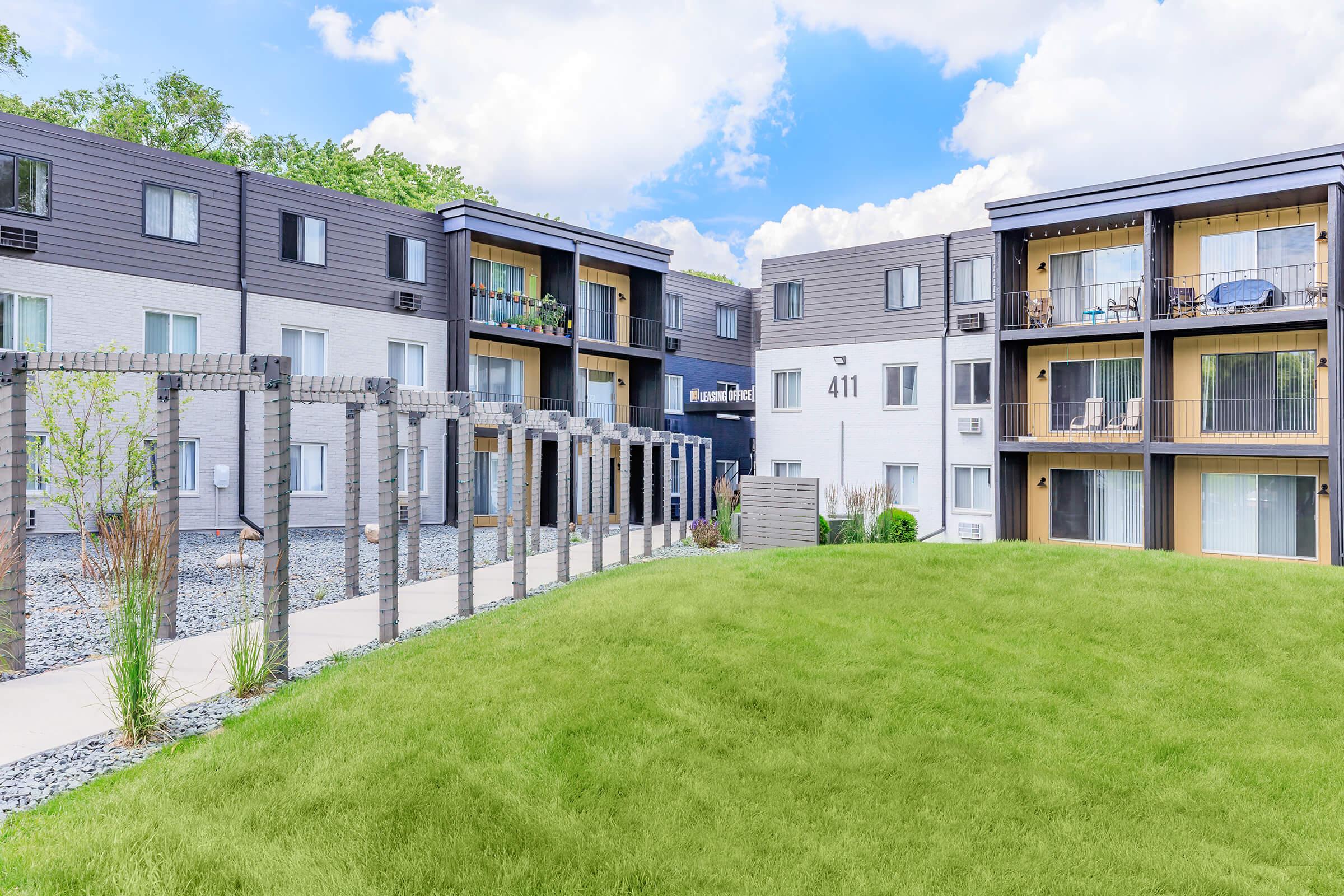 A modern apartment complex with two separate buildings featuring balconies. The foreground includes a well-maintained green lawn and a pathway leading to the entrance. Decorative columns and landscaping enhance the outdoor space, with clouds visible in the blue sky above.