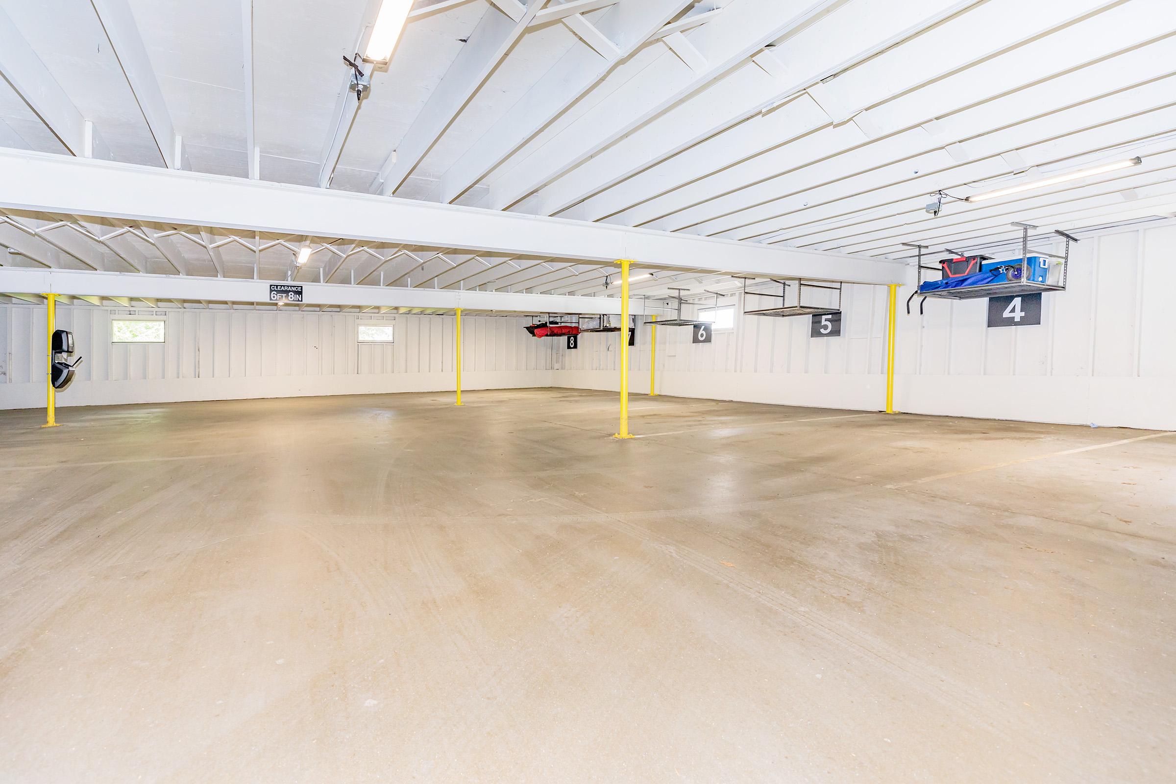 A spacious, empty garage with white walls and a light-colored floor. It features several yellow support poles and overhead storage. There are numbered parking spaces marked on the walls, with some visible wall mounts for bikes or storage. Natural light comes in through a couple of windows.