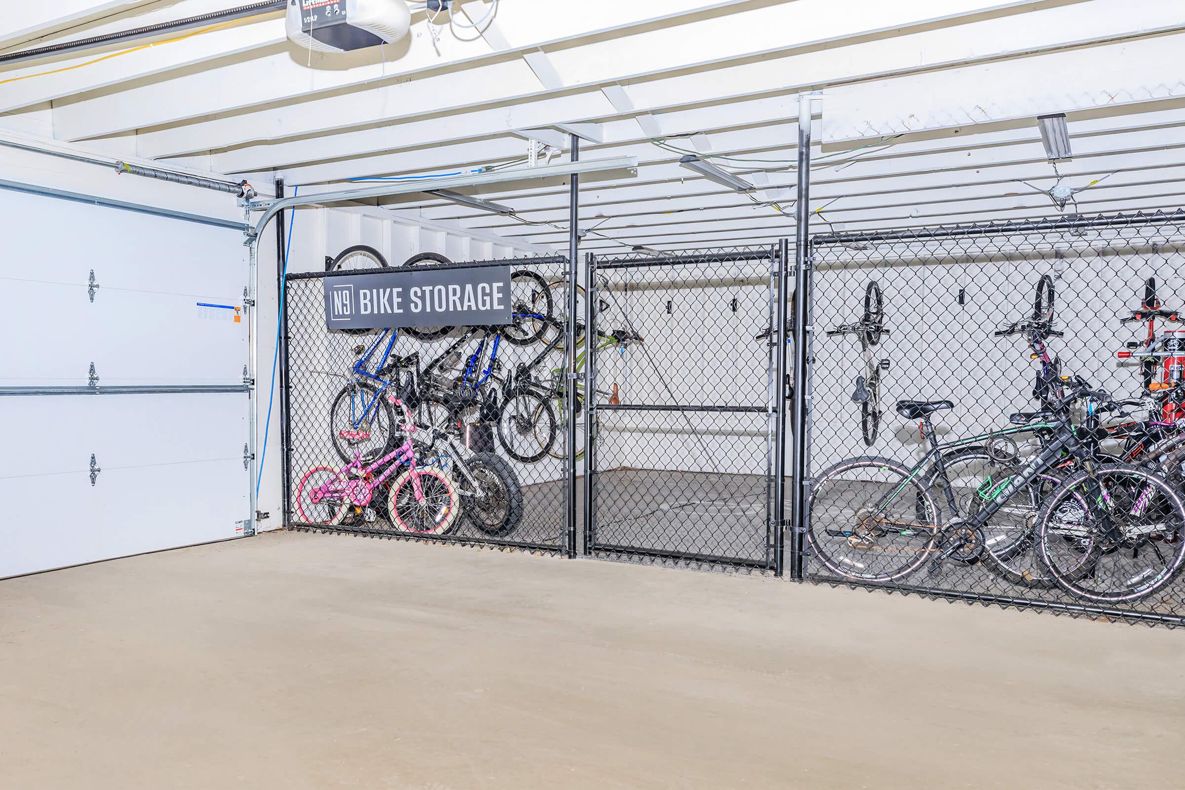 A clean garage space featuring a chain-link fence enclosure labeled "Bike Storage." Inside, several bicycles of various colors and types are neatly hung and secured. The garage has a concrete floor and an overhead door, providing a bright and organized area for bike storage.