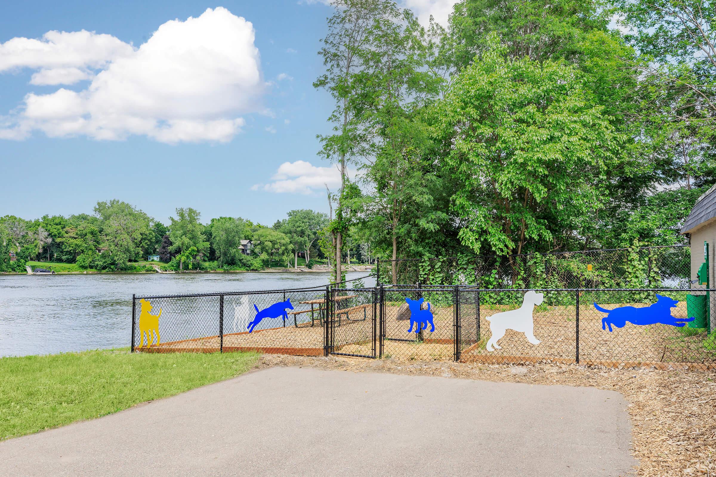 A fenced dog park near a river, featuring colorful silhouettes of dogs in blue, yellow, and white on the fence. The park is surrounded by lush greenery, and a paved path leads towards it. The sky is clear with a few clouds, creating a bright and inviting atmosphere for pets and their owners.