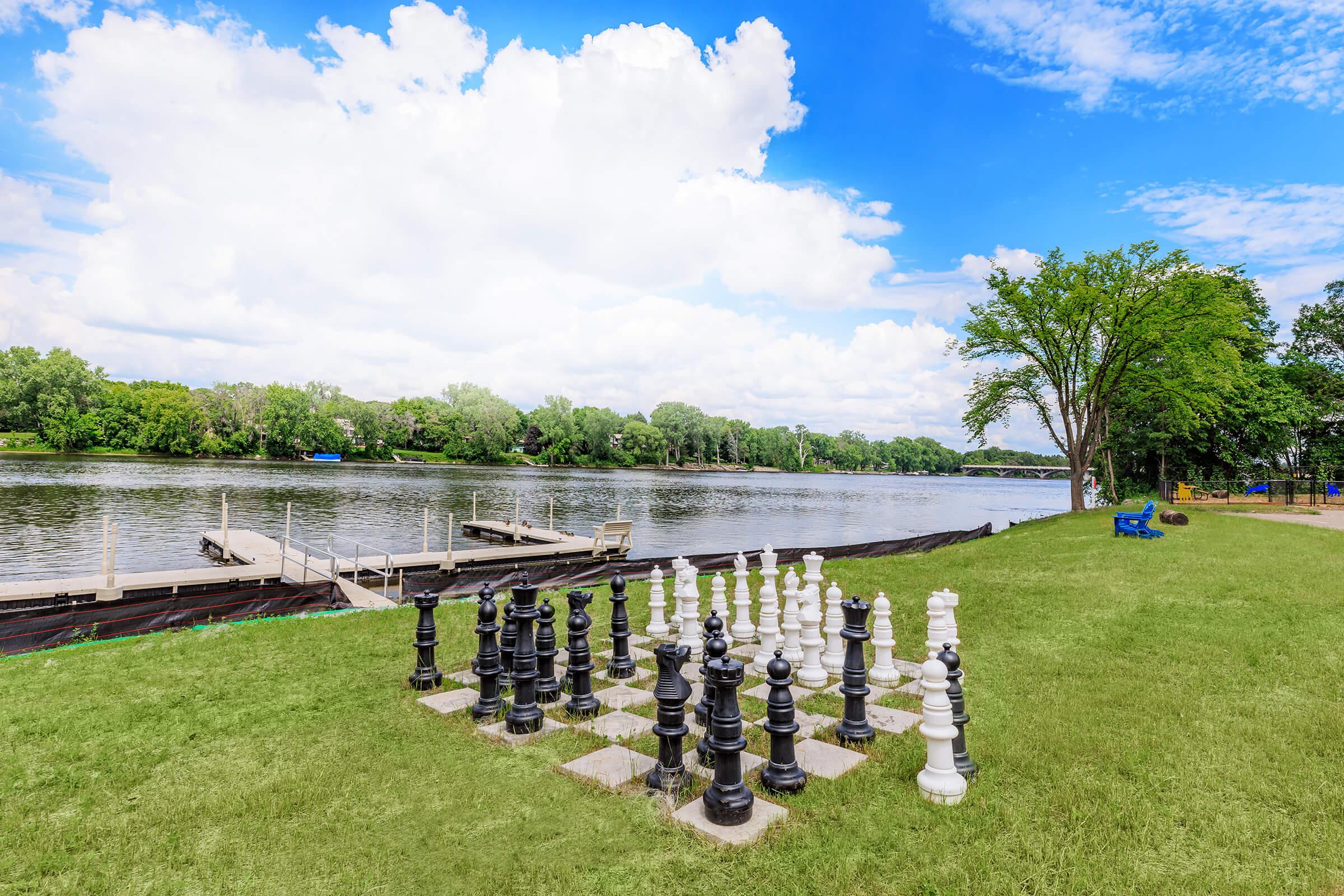 A large outdoor chess set made of black and white pieces is situated on a grassy area near a river. In the background, trees line the riverbank under a bright blue sky with scattered clouds. A dock and seating area are visible along the river.