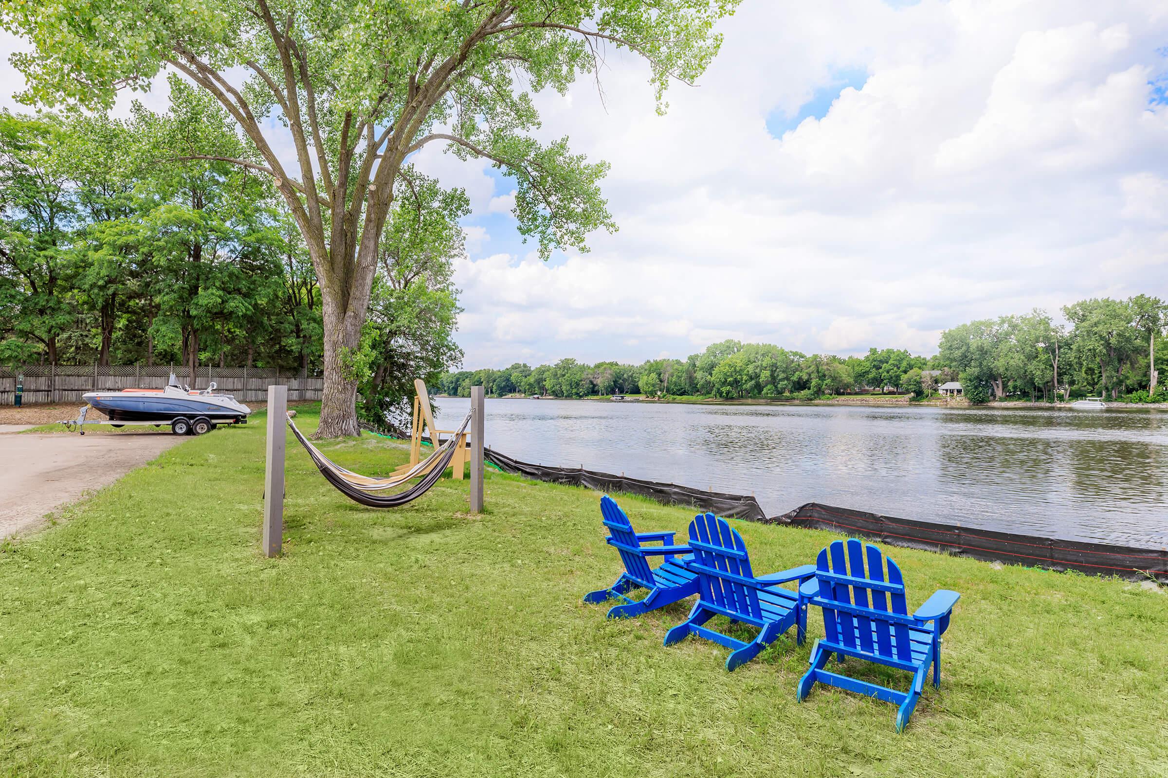 A serene lakeside view featuring two blue Adirondack chairs facing the water, with a hammock hanging nearby. In the background, a boat is parked on the shore, and lush green trees surround the area under a partly cloudy sky. The scene captures a peaceful outdoor retreat.