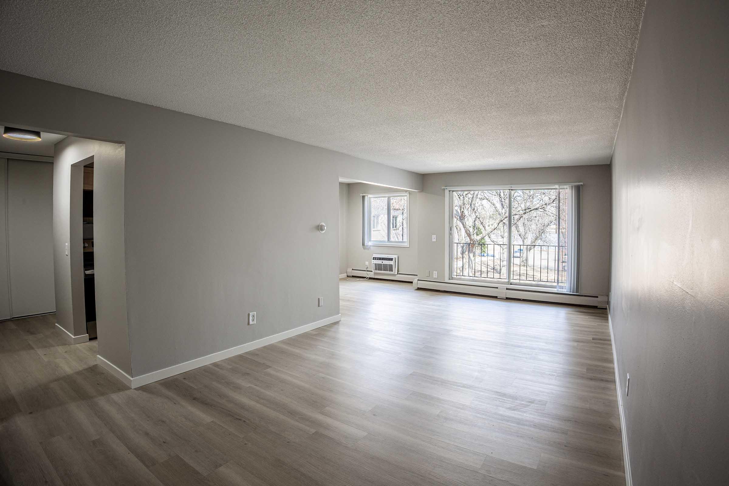 Spacious, empty living area with light-colored walls and wooden flooring. Large windows allowing natural light, with a view of trees outside. Minimalistic design and neutral tones create a bright and open atmosphere. An adjacent wall features an entrance to a separate room or area.