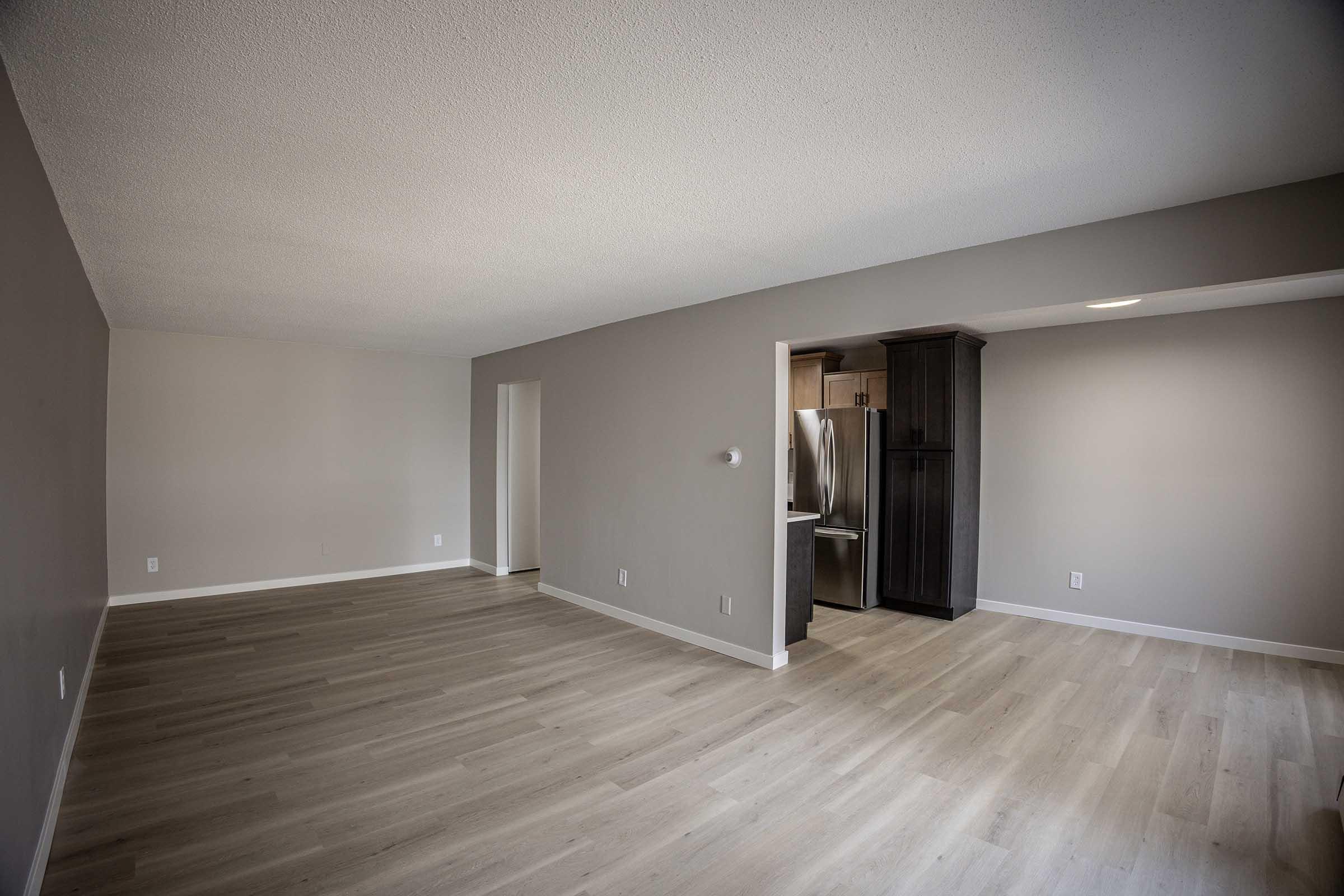 A spacious, empty living room with light gray walls and a smooth wooden floor. To the right, there’s an open doorway leading to a kitchen area featuring stainless steel appliances. The room is well-lit with natural light coming in, creating a bright and airy atmosphere.