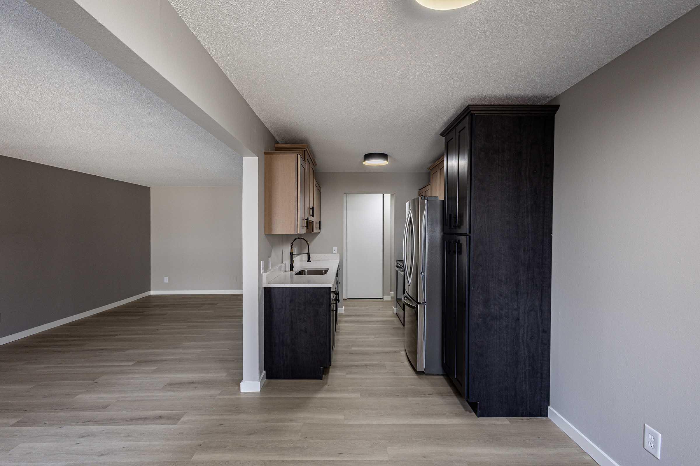 A modern kitchen in an open-concept living space features dark cabinetry, a stainless steel refrigerator, and a sink. The floor is light-colored wood, and the walls are painted in neutral tones. The area is well-lit with natural light coming from adjacent rooms.