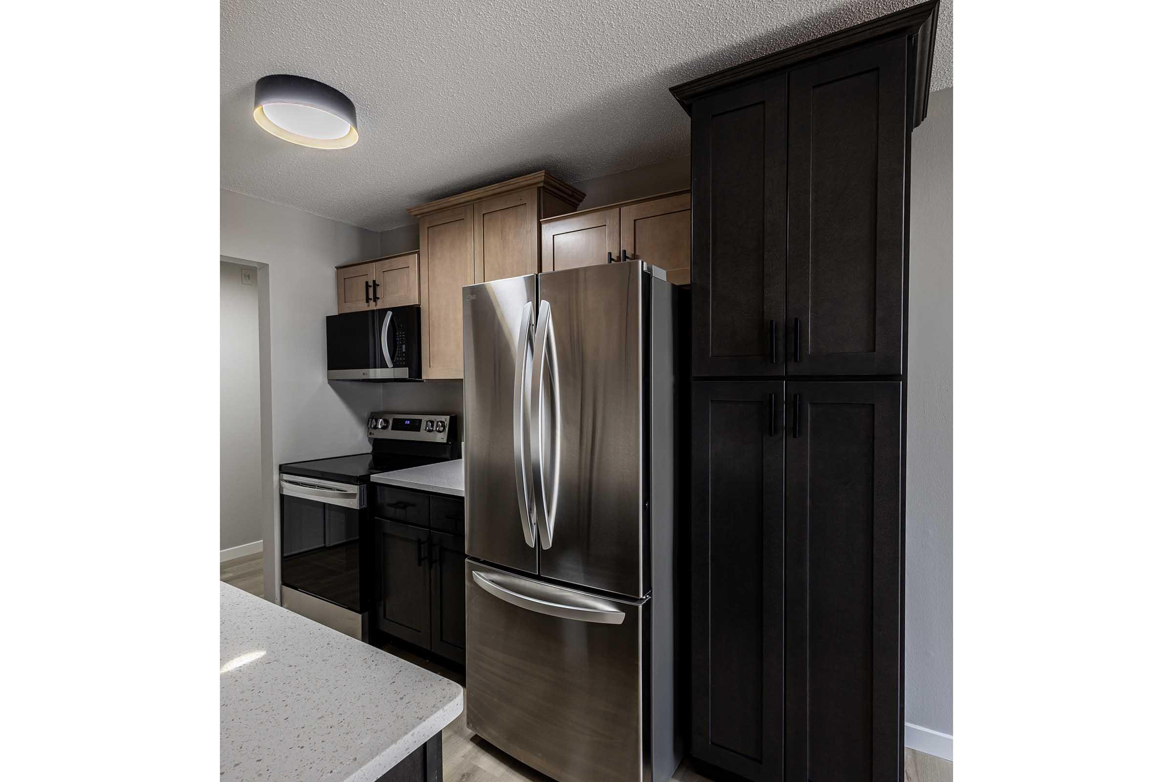 Modern kitchen featuring stainless steel refrigerator and microwave, black appliances, light countertops, and a blend of dark and light wooden cabinets. A round ceiling light illuminates the space, showcasing the contemporary design.