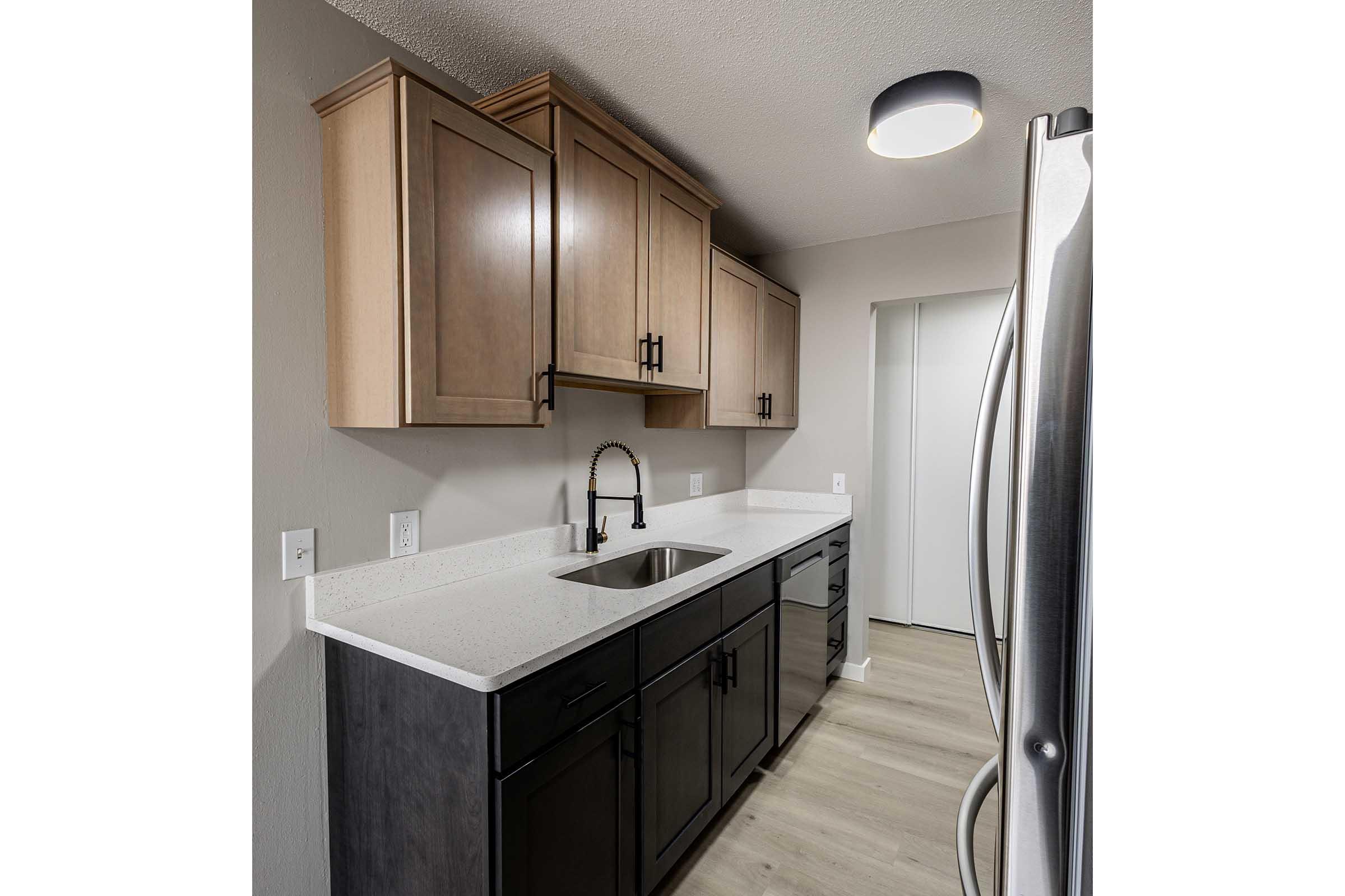 Modern kitchen featuring light wood cabinetry and dark lower cabinets, a white countertop, and a sleek black faucet at the sink. Stainless steel refrigerator and a ceiling light above. Neutral wall color and flooring contribute to a clean and contemporary look. Door on the right leads to another room.