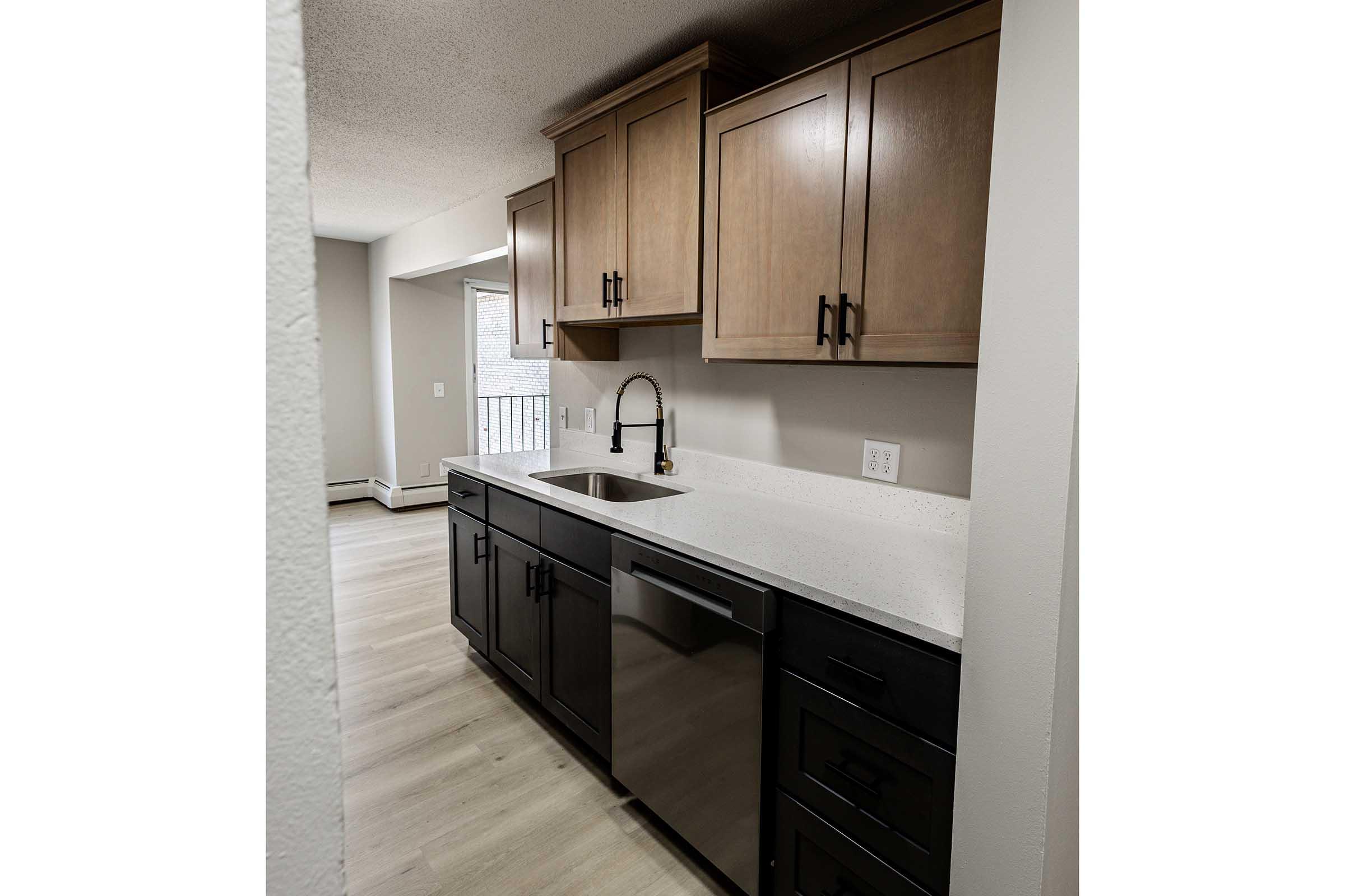 Modern kitchen featuring light wood cabinets, dark lower cabinetry, and a stainless steel dishwasher. The countertop is a light, speckled stone. A faucet is installed above a single-basin sink. The kitchen is well-lit, with a large window allowing natural light. The flooring is light-colored laminate.