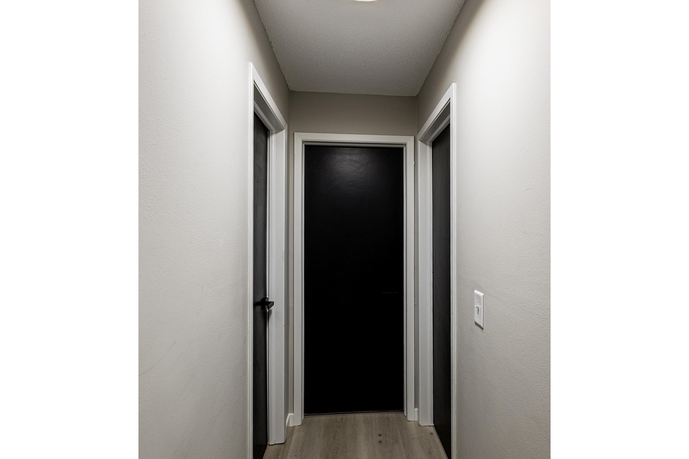 Narrow hallway with light gray walls and wood flooring. Two doors on either side, one leading to a dark-colored door at the end of the hallway. The ceiling is illuminated by a light fixture, creating a bright and clean space.