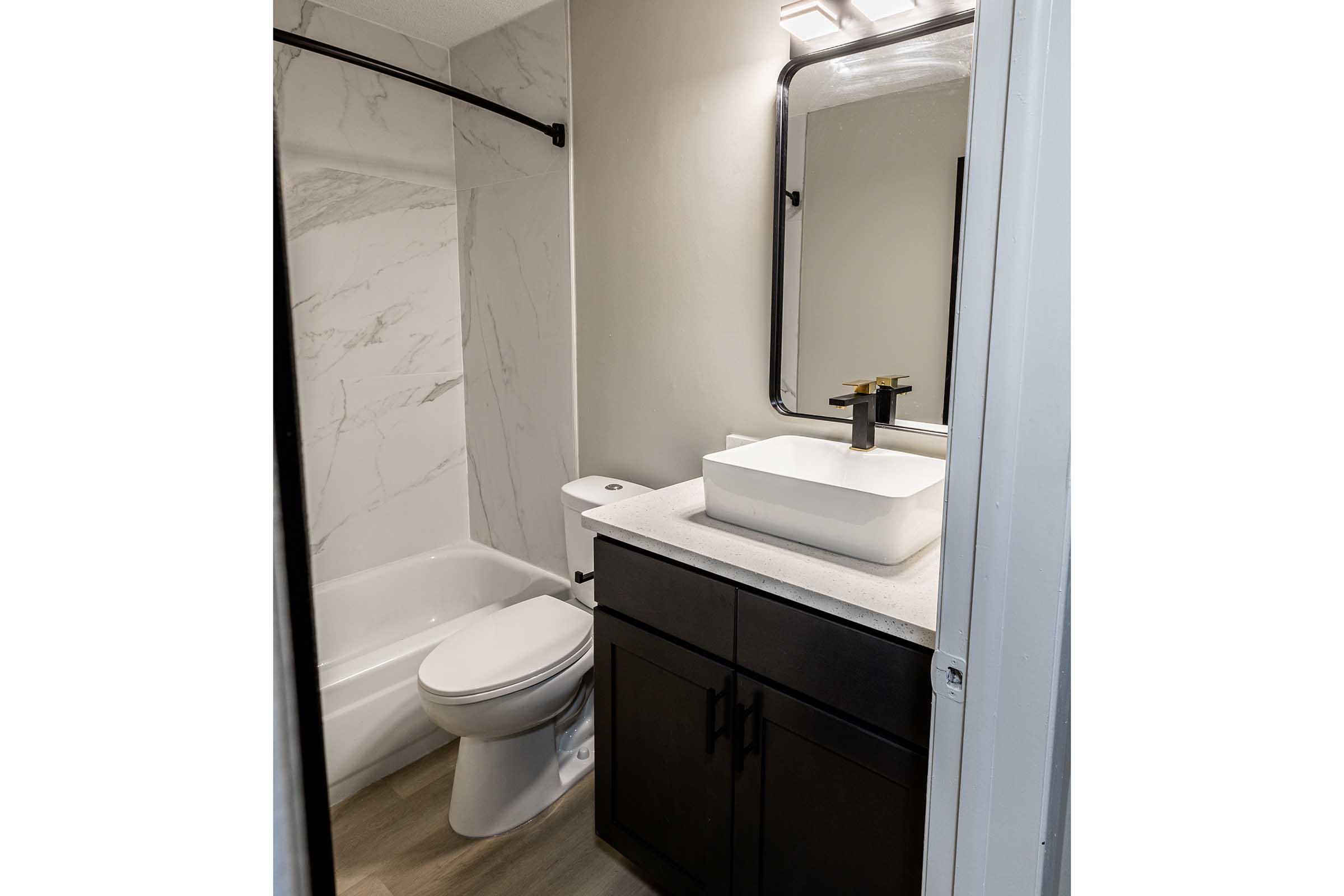 A modern bathroom featuring a walk-in shower with white marble walls, a sleek black cabinet with a white vessel sink on a countertop, a modern faucet, a toilet, and a large mirror above the sink. The walls are painted in a neutral tone, and the flooring is light-colored laminate.