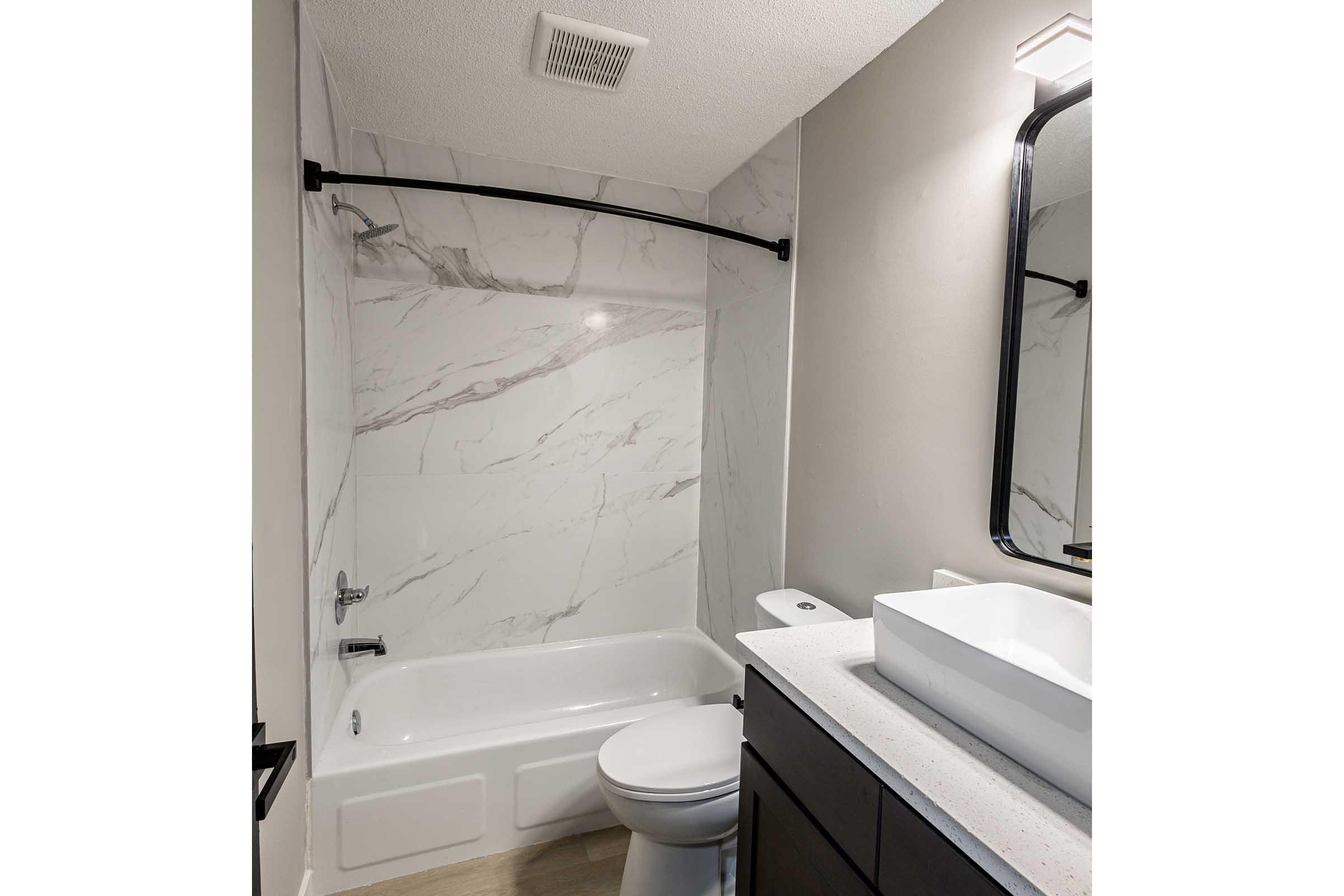 A modern bathroom featuring a white tub with a marble surround, a wall-mounted mirror above a sleek sink, and dark cabinetry. The walls are painted gray, and the space is illuminated by a wall sconce. The overall design is clean and contemporary, enhancing the bathroom's aesthetics.