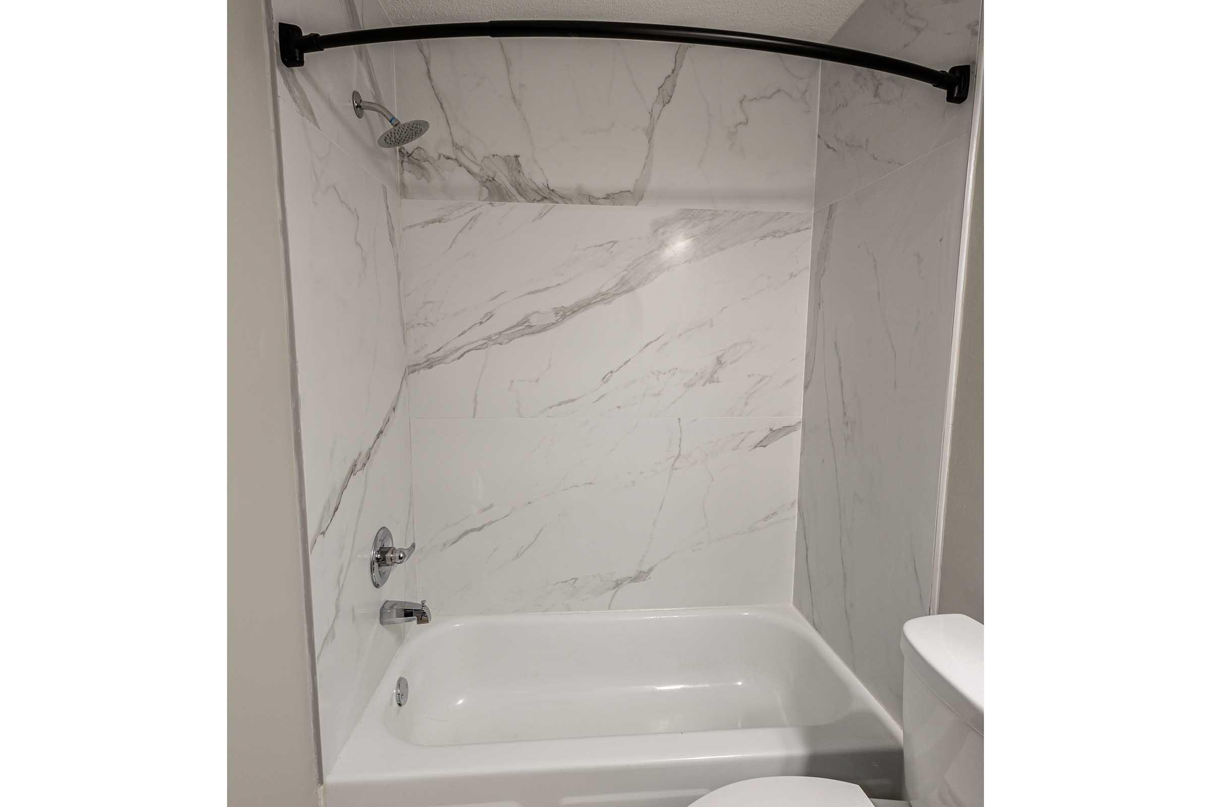 A modern bathroom featuring a bathtub with a showerhead above. The walls are covered in large, white marble tiles. The bathtub is white and the design is sleek and minimalistic. There is a toilet visible on the right side of the image. The overall aesthetic is clean and contemporary.