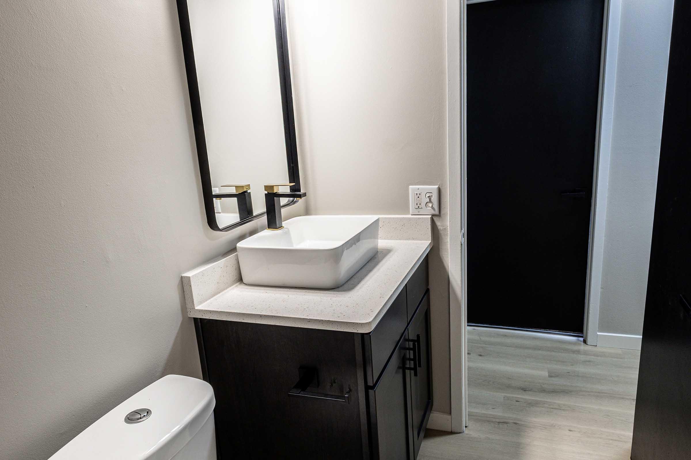 A modern bathroom featuring a white square sink on a light countertop, dark cabinetry, and two mirrors above the sink. A black door is visible in the background, while a light wood floor adds warmth to the space. The walls are painted in a neutral tone, creating a sleek and contemporary atmosphere.