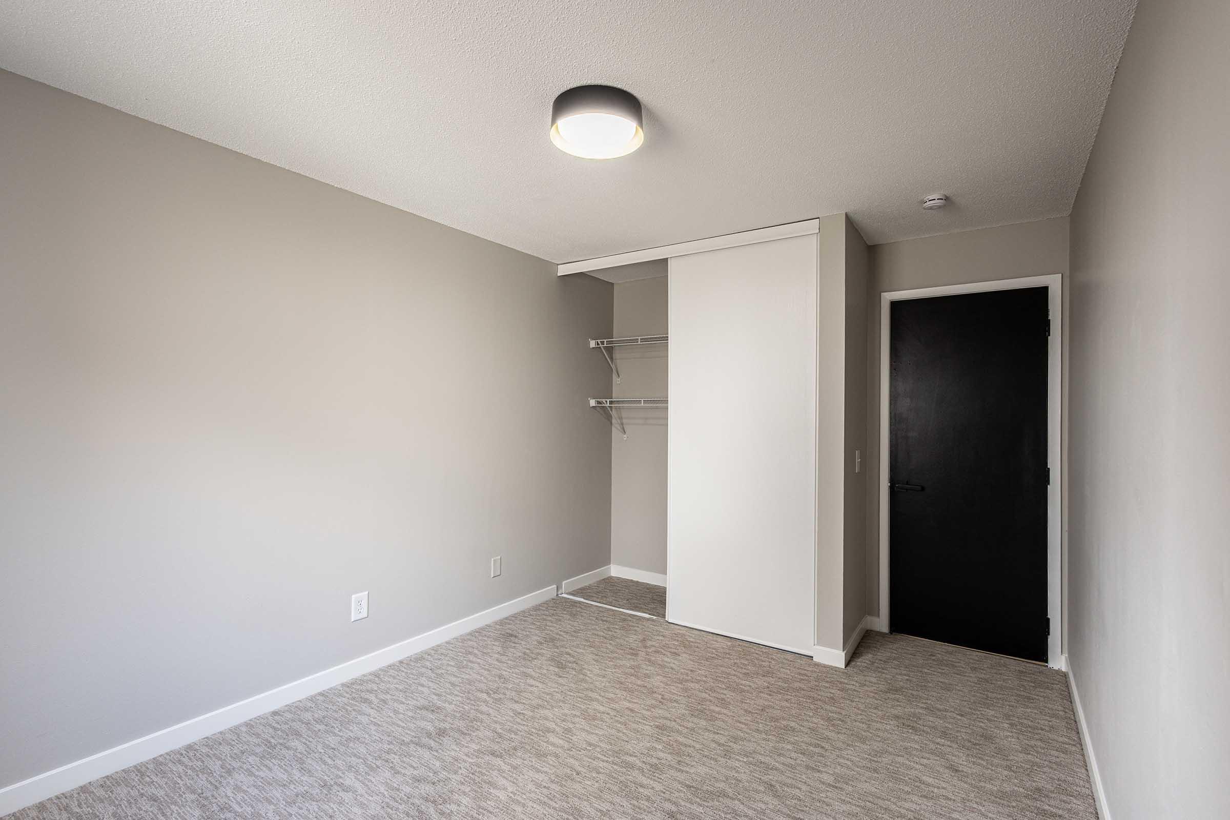 A vacant room featuring light gray walls and a carpeted floor. On one side, there is a closet with metal shelving, and a black door on the opposite side. A modern ceiling light fixture illuminates the space.