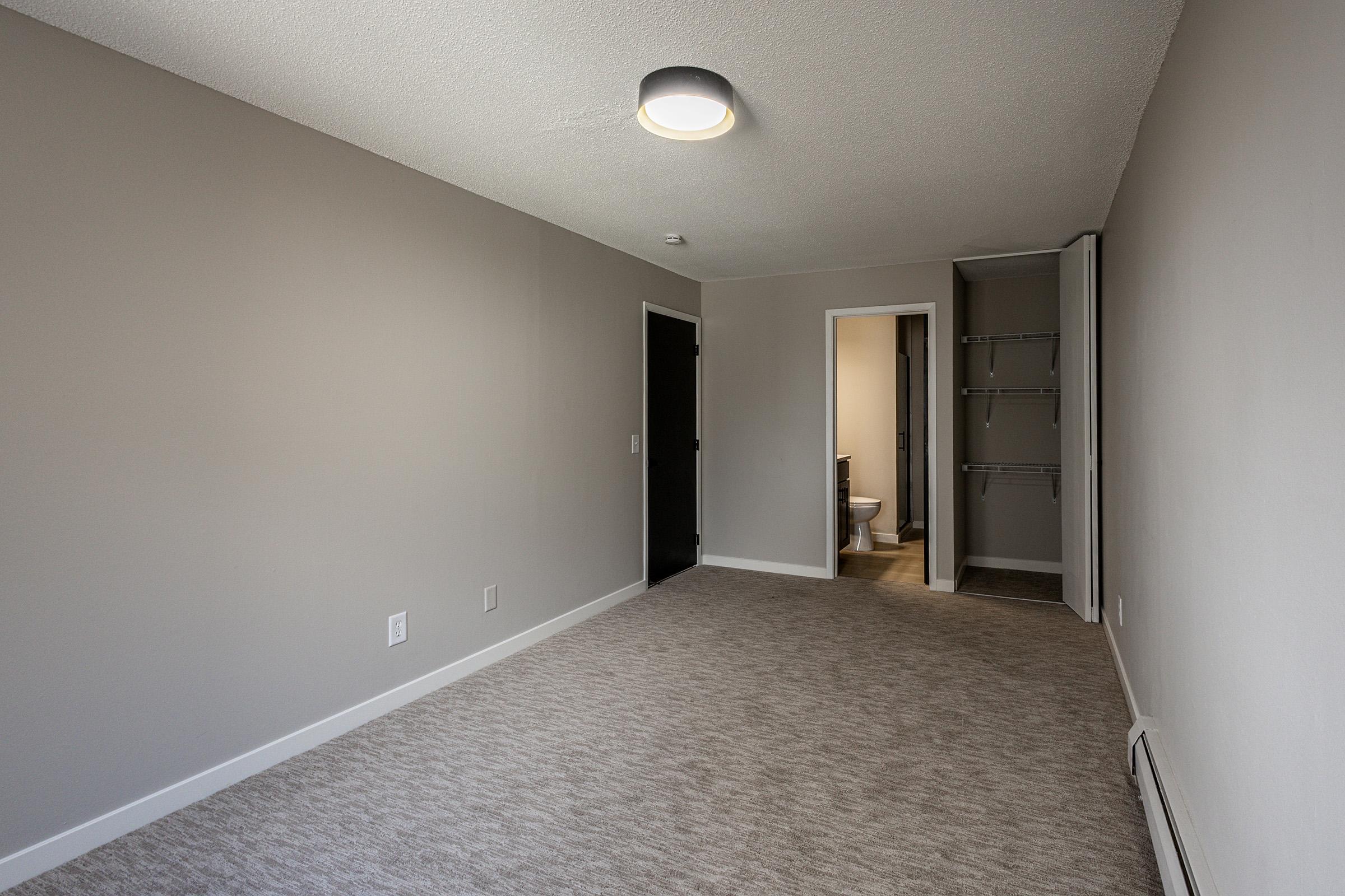 A spacious, freshly painted room with light gray walls and carpeted flooring. The room features a ceiling light and a doorway leading to a bathroom. A closet with shelves is visible on one side, while the opposite wall has a solid black door. Natural light enhances the bright and airy ambiance.