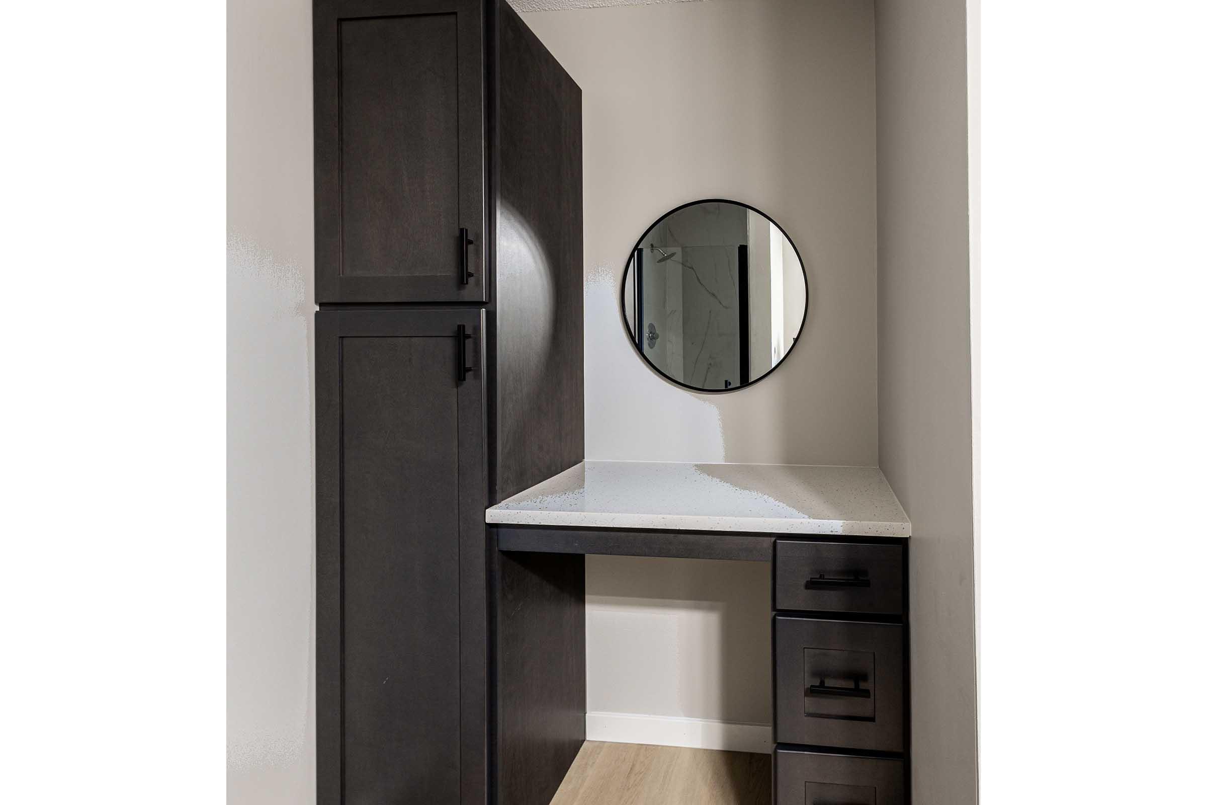 A modern, minimalist corner of a room featuring a dark wood cabinet, a circular mirror on the wall, and a sleek white countertop with storage drawers. The walls are painted a light color, lending a bright and airy feel to the space.