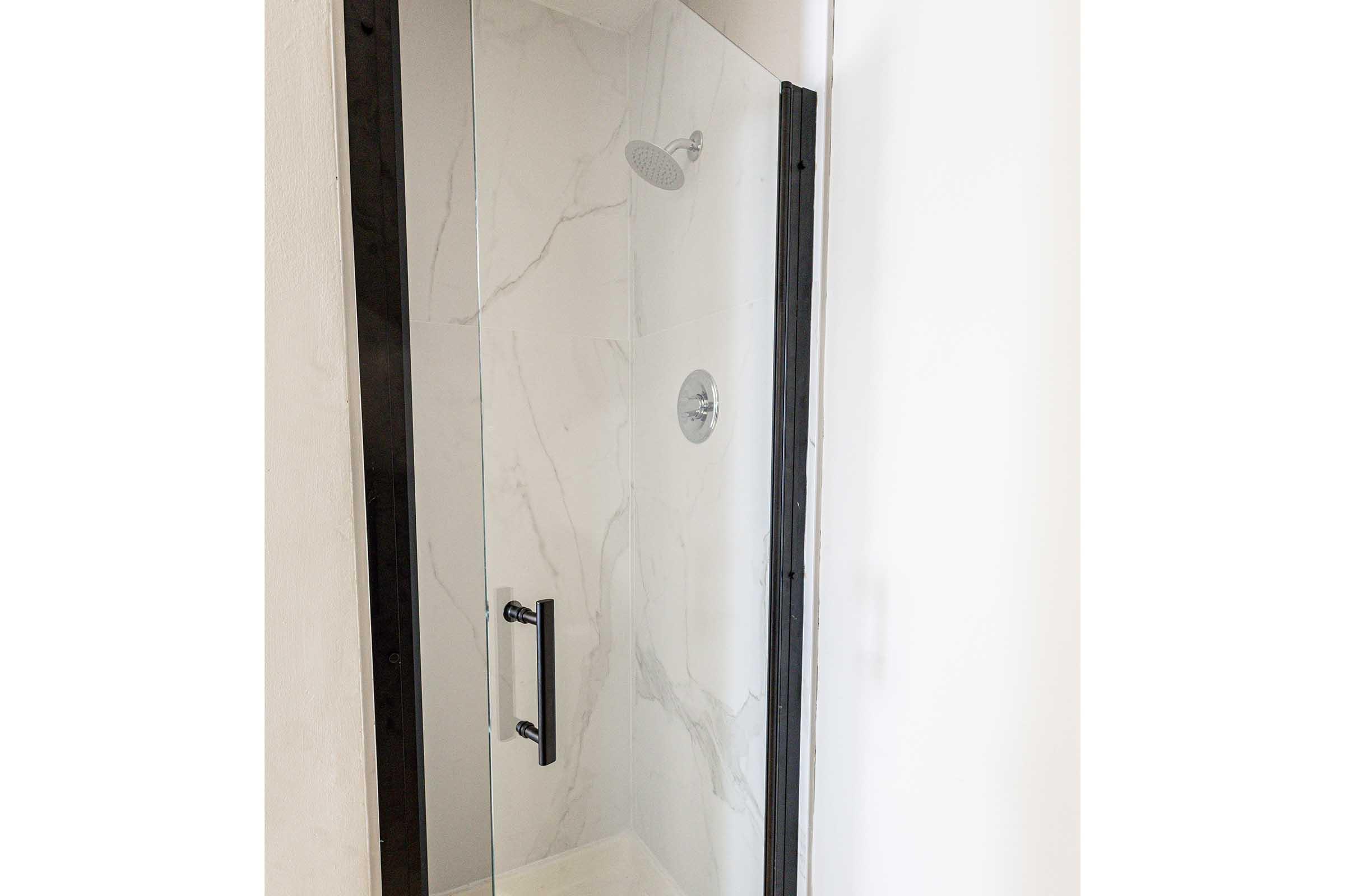 A modern glass shower enclosure featuring a marble-like wall design. The enclosure includes a sleek handle and a rain showerhead mounted on the ceiling. The interior is clean and minimalist, with bright lighting and white accents.