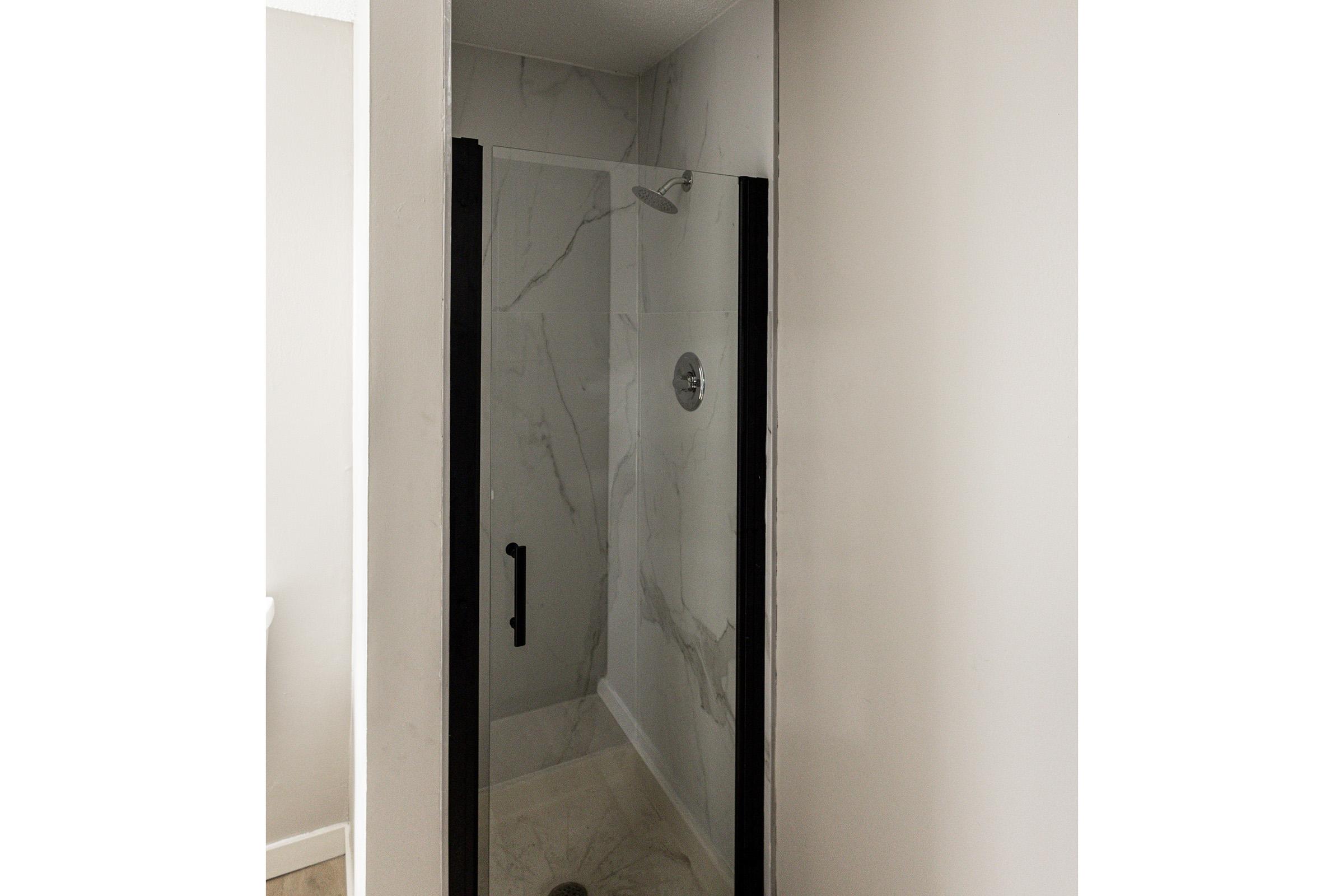 A modern, glass-enclosed shower with black framing, featuring a rainfall showerhead and marble-like tiled walls. The interior is well-lit and spacious, showcasing a minimalist design within a bright bathroom.