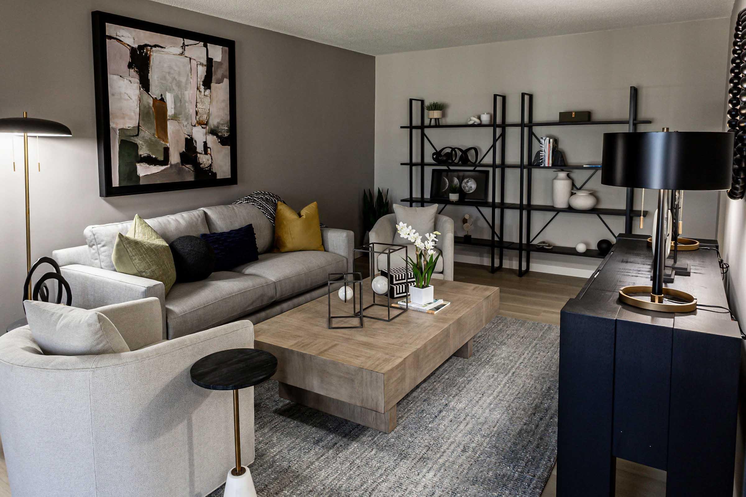 A modern living room featuring a light gray sofa with decorative pillows, a large wooden coffee table, and a stylish black console. There’s an abstract painting on the wall, a floor lamp, and a few indoor plants. A minimalist bookshelf displays various decorative items, creating a cozy and contemporary atmosphere.