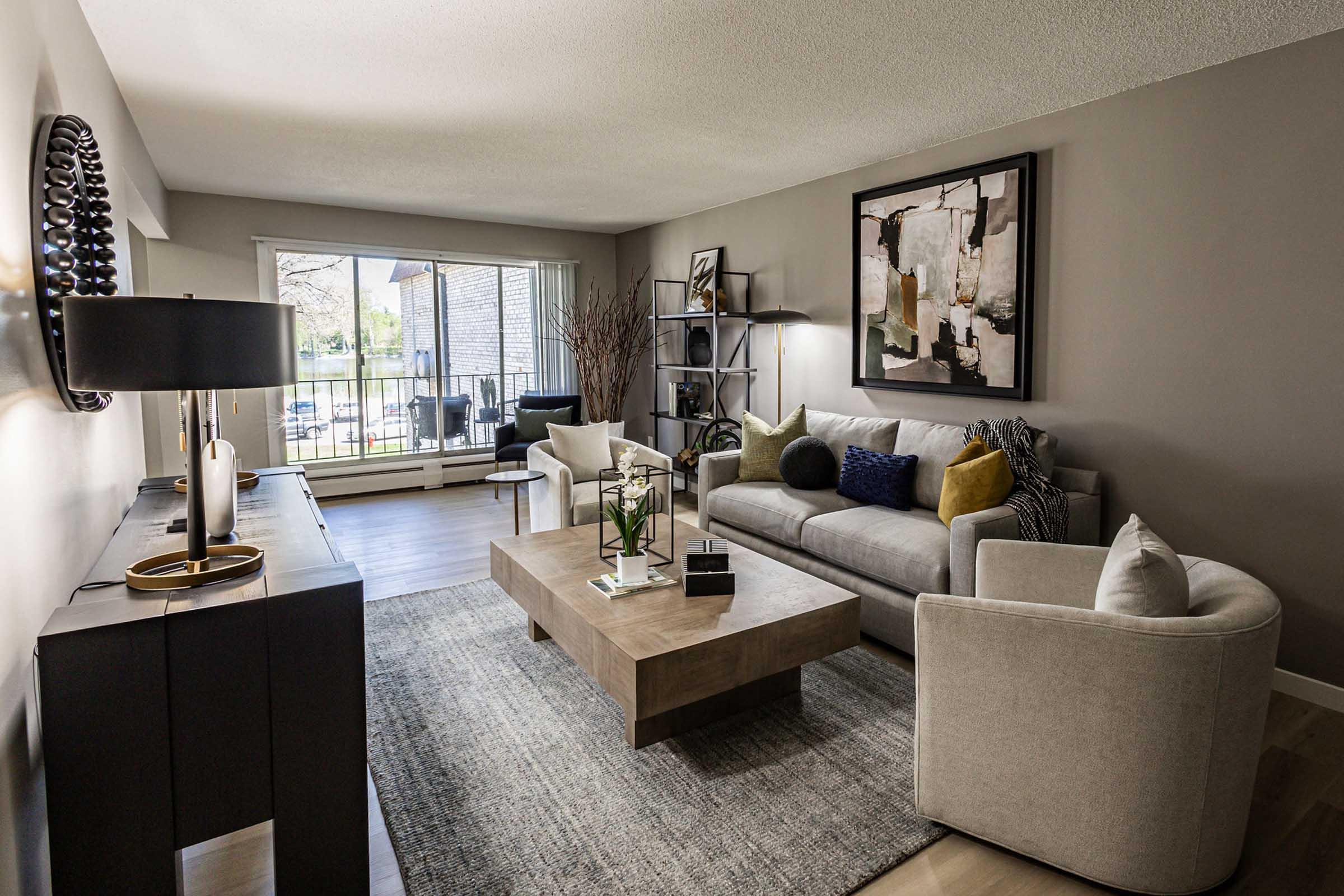 A modern living room featuring a light gray sofa with colorful cushions, a wooden coffee table, and two armchairs. A stylish lamp and decorative items are on a dark console table. Large windows provide natural light, revealing an outdoor view. Neutral wall colors create a warm and inviting atmosphere.