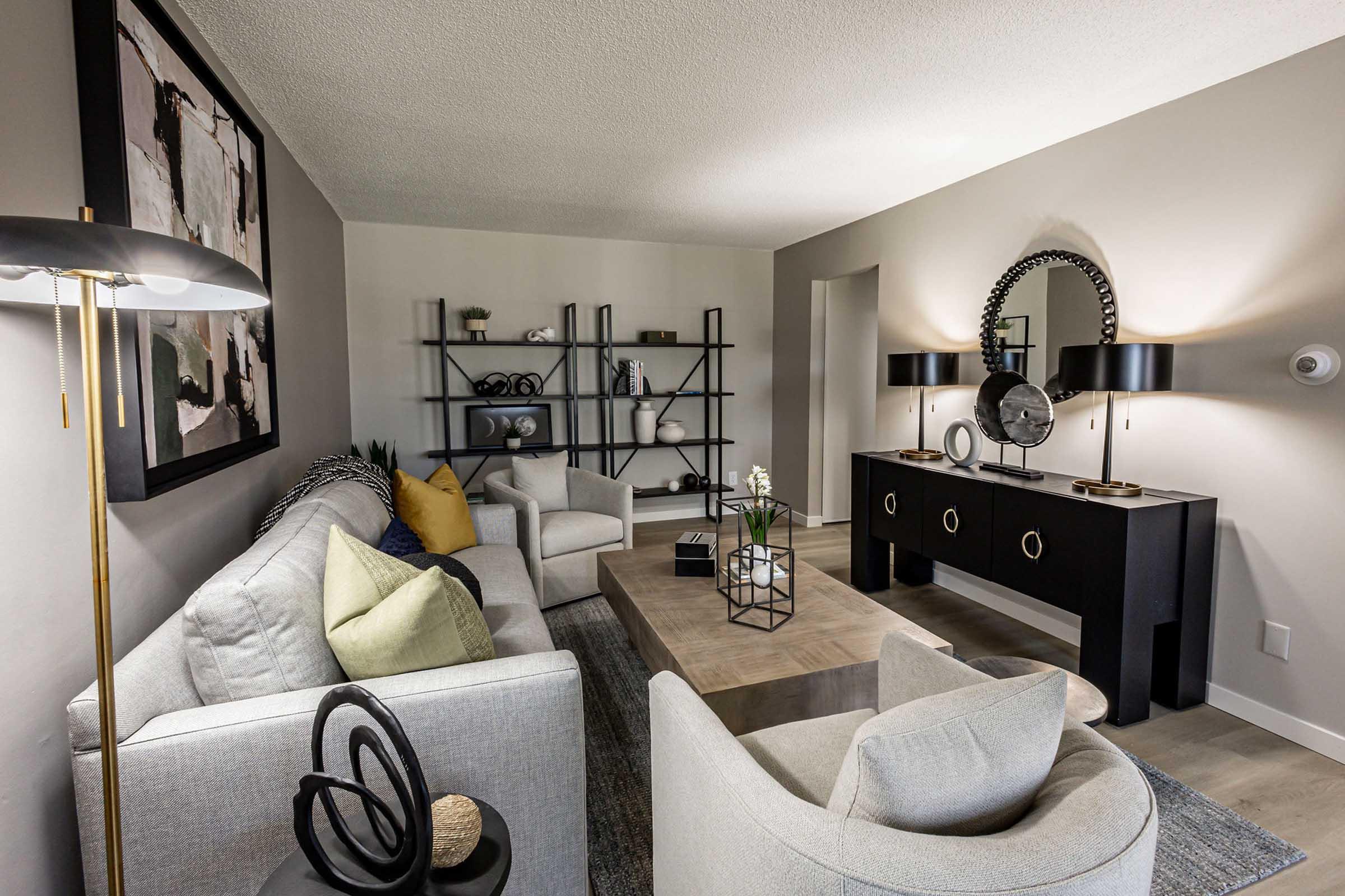 A modern living room featuring a light gray sofa and two armchairs with decorative pillows, a coffee table, and a black console table. The wall displays abstract artwork, and there's a decorative bookshelf with vases and plants. Soft lighting from floor lamps adds a warm ambiance.