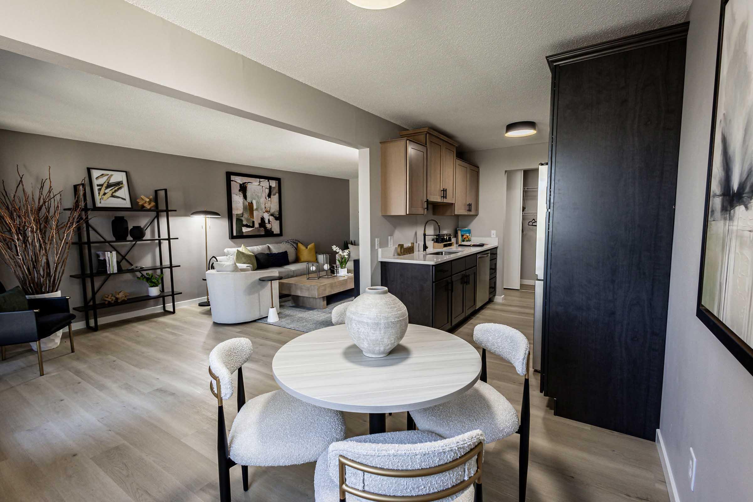 Modern interior of a spacious living area featuring a dining table with a decorative vase, a cozy seating area with a sofa and armchairs, a stylish kitchen with wooden cabinetry, and contemporary decor. Natural light flows through large windows, creating a welcoming atmosphere.