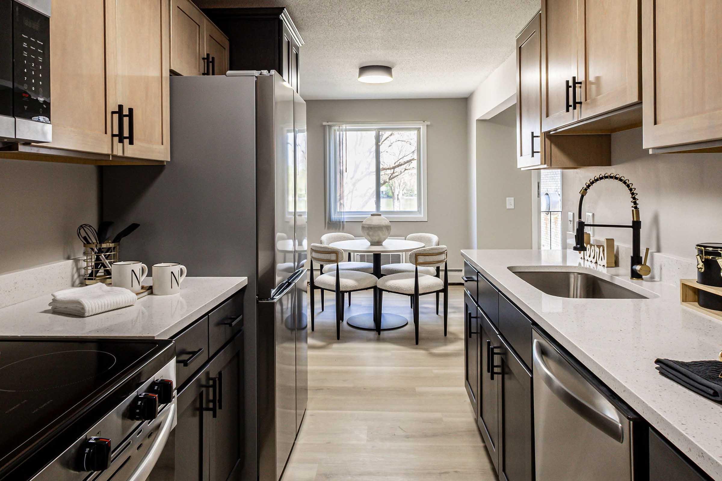 Modern kitchen featuring stainless steel appliances, including a fridge and stove, with light wood cabinetry and white countertops. A dining area with a round table and upholstered chairs is visible through a window, illuminated by natural light. The overall design is sleek and contemporary.