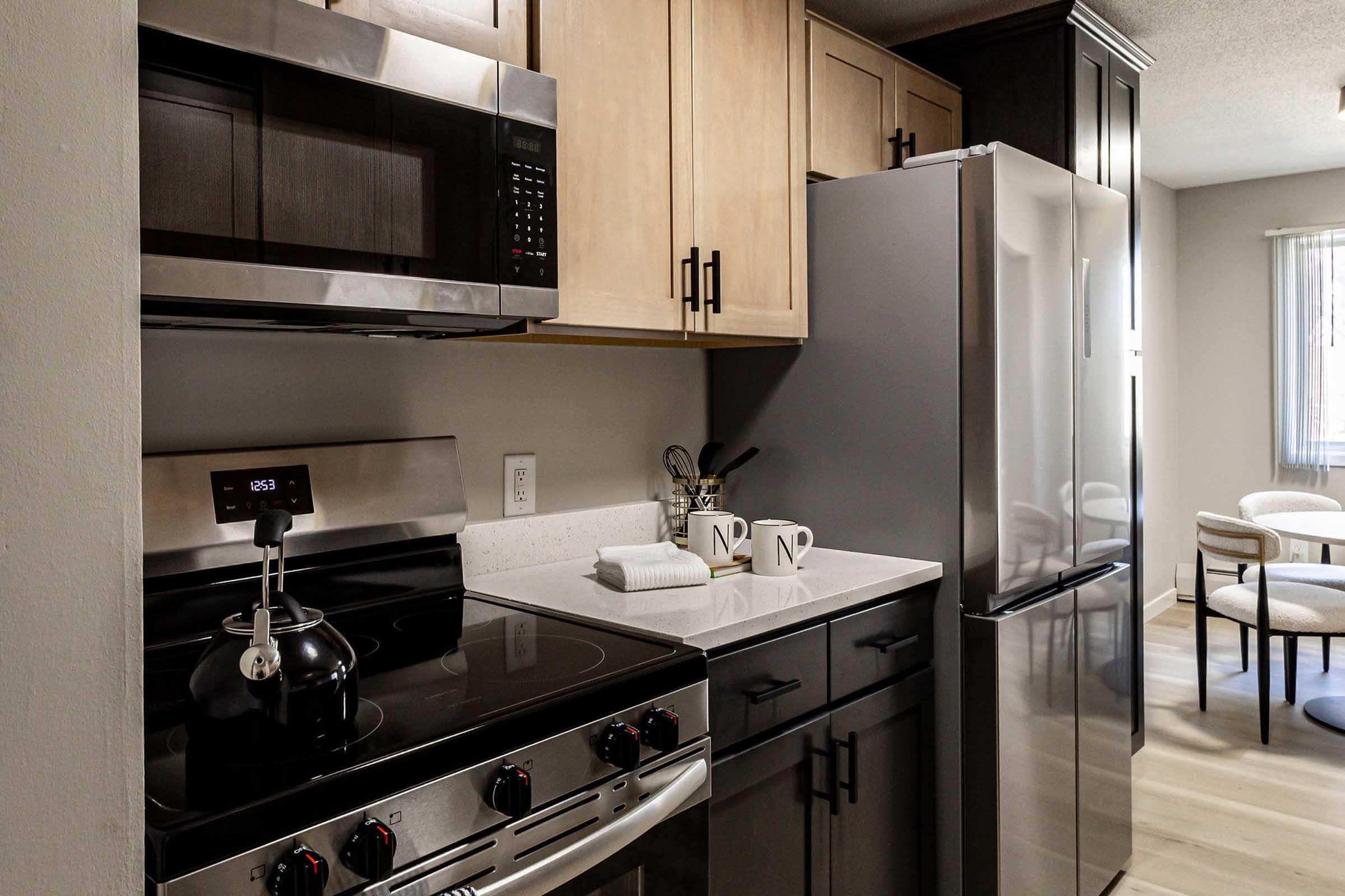 A modern kitchen featuring stainless steel appliances, including a microwave and refrigerator. The cabinetry has a mix of light and dark wood finishes. A kettle is on the stove, and two mugs are neatly placed on the countertop. The space is well-lit with natural light coming from a nearby window.