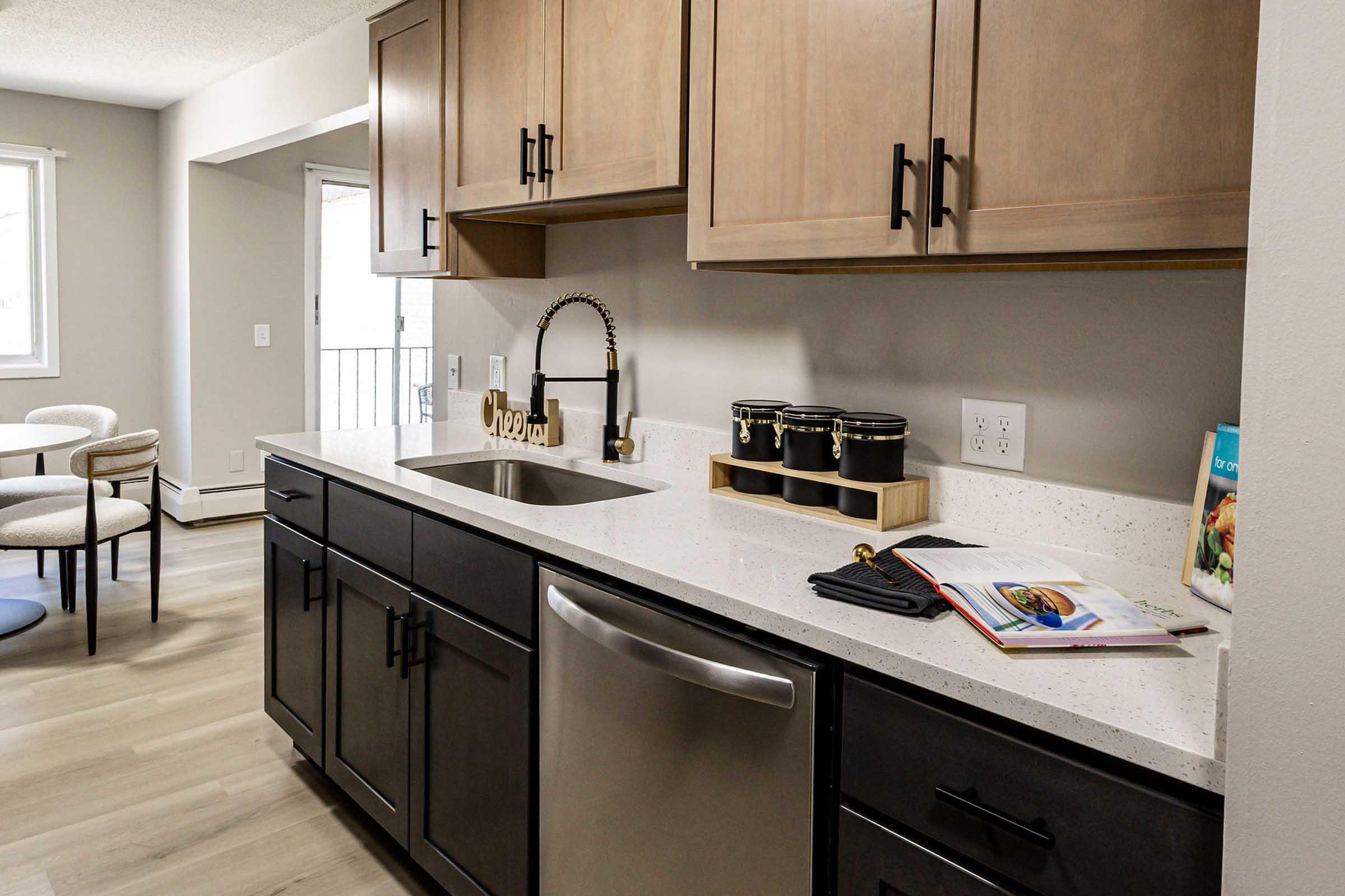 A modern kitchen with wooden cabinetry, a stainless steel sink, and a dishwasher. The countertop is light-colored with a few decorative items, including jars and a cookbook. A small dining area with a round table and chairs is visible in the background, with natural light coming from a window.