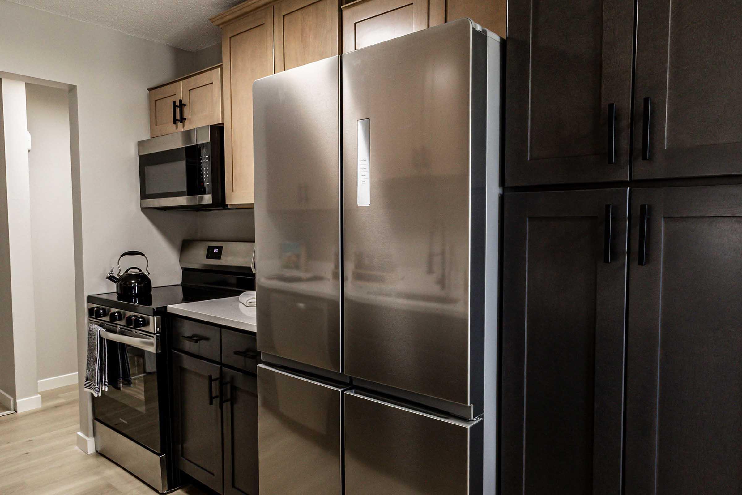Modern kitchen featuring stainless steel appliances, including a double-door refrigerator, microwave, and stove. The cabinetry is a mix of light wood and dark finishes, with a black countertop. A kettle is placed on the stove, and towels hang from the oven handle. Bright, neutral walls complement the overall design.