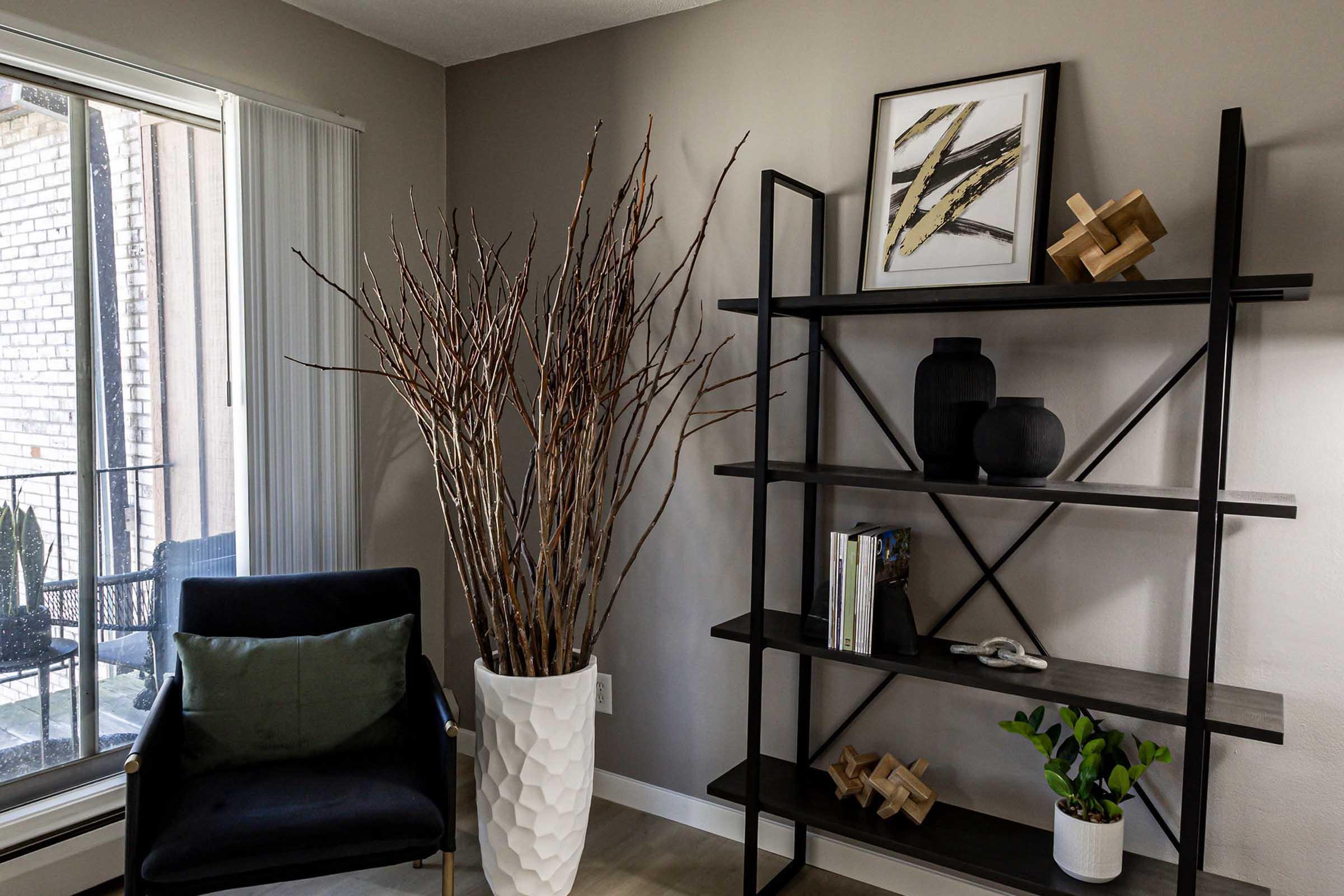 A modern interior space featuring a tall white planter with branches, a black open-shelf unit displaying decorative items, a black vase, and a small plant. There's a comfortable chair in the corner and large windows letting in natural light.