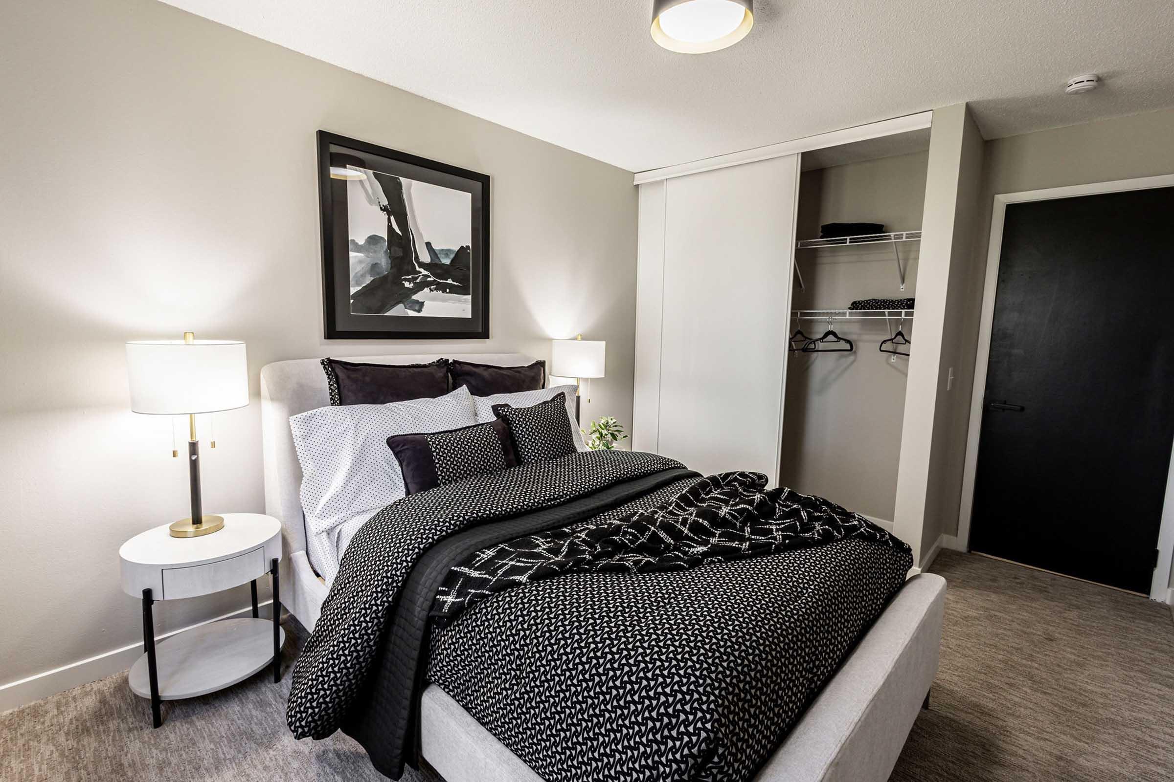 A modern bedroom featuring a neatly made bed with black and white bedding, two decorative pillows, and a bedside table with a lamp. A framed artwork is hung on the wall above the bed, and there is a closet with hangers visible in the background. The overall decor is minimalist and stylish.