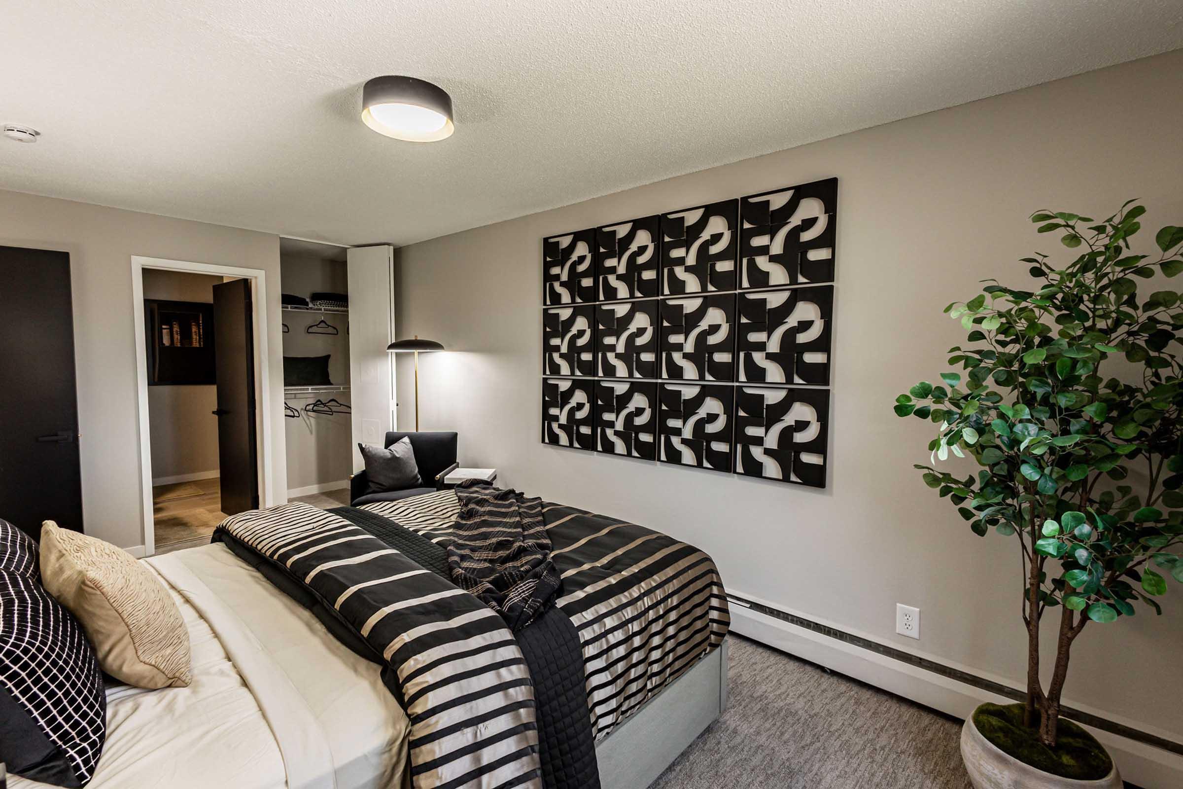 A modern bedroom featuring a neatly made bed with black and white striped bedding. There's a decorative wall art piece above the bed, a green indoor plant in one corner, and a sleek floor lamp next to it. A closet is visible in the background, with a light-colored door leading to another room.