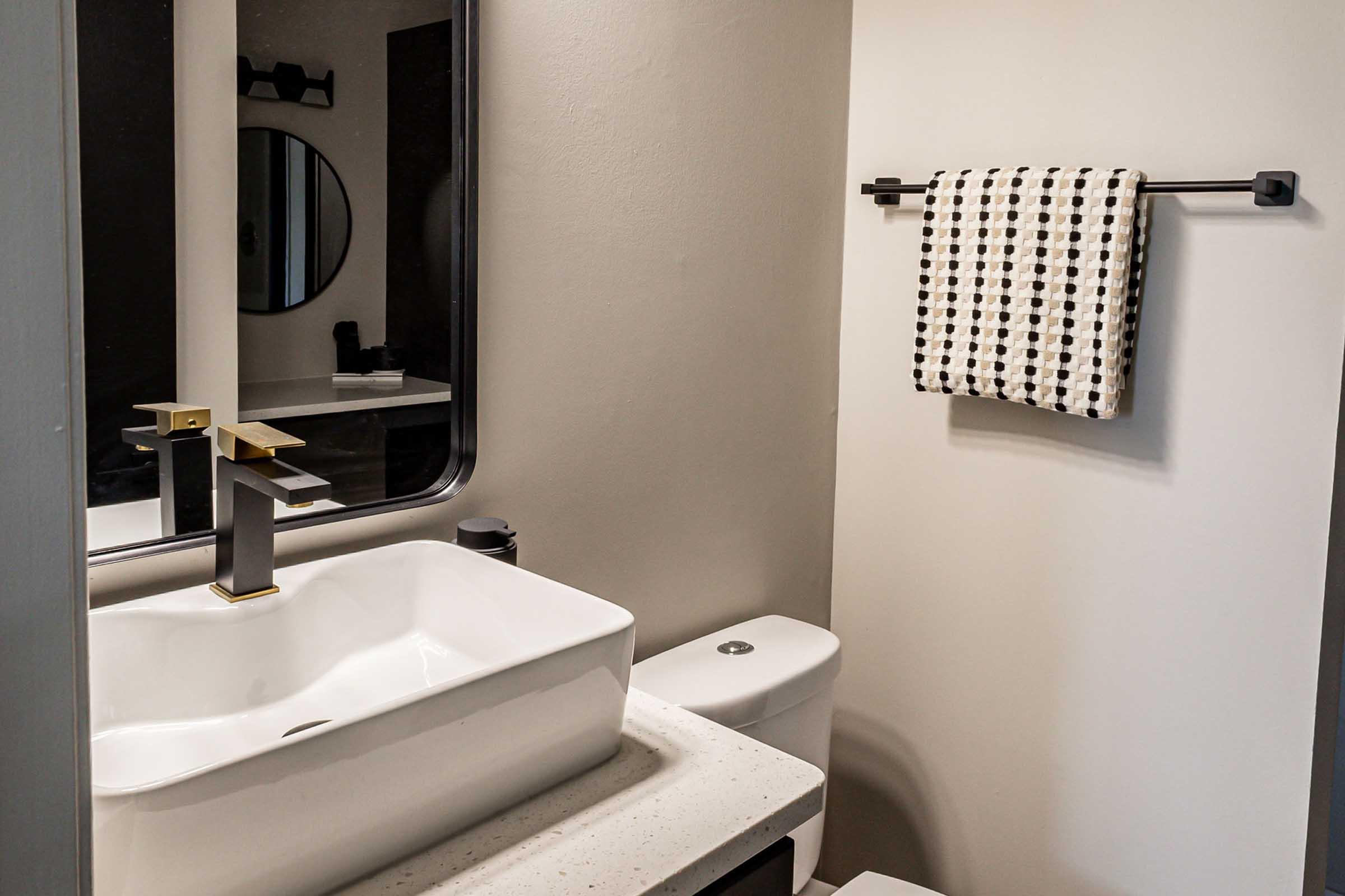 Modern bathroom interior featuring a sleek, rectangular sink with a gold faucet, a round mirror above, and a towel rack with a patterned black and white towel. The walls are painted in neutral tones, and there's a compact toilet visible. A minimalistic design emphasizes cleanliness and functionality.