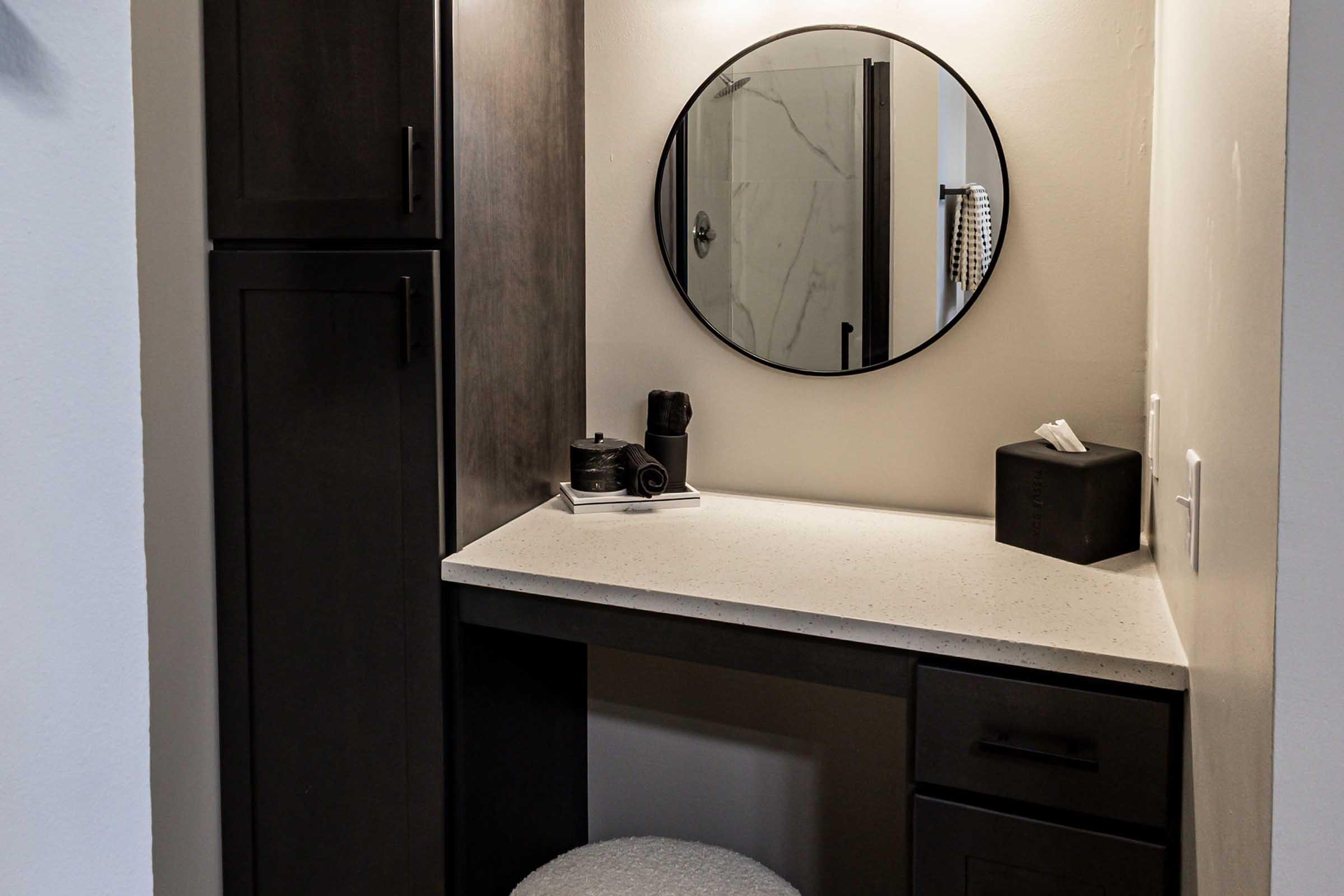 A modern bathroom nook featuring a round mirror mounted on a light-colored wall. There is a dark wooden cabinet to the left and a light-colored countertop with a small black box, a tissue box, and a camera. A fluffy stool is positioned in front of the counter, creating a stylish grooming or working space.