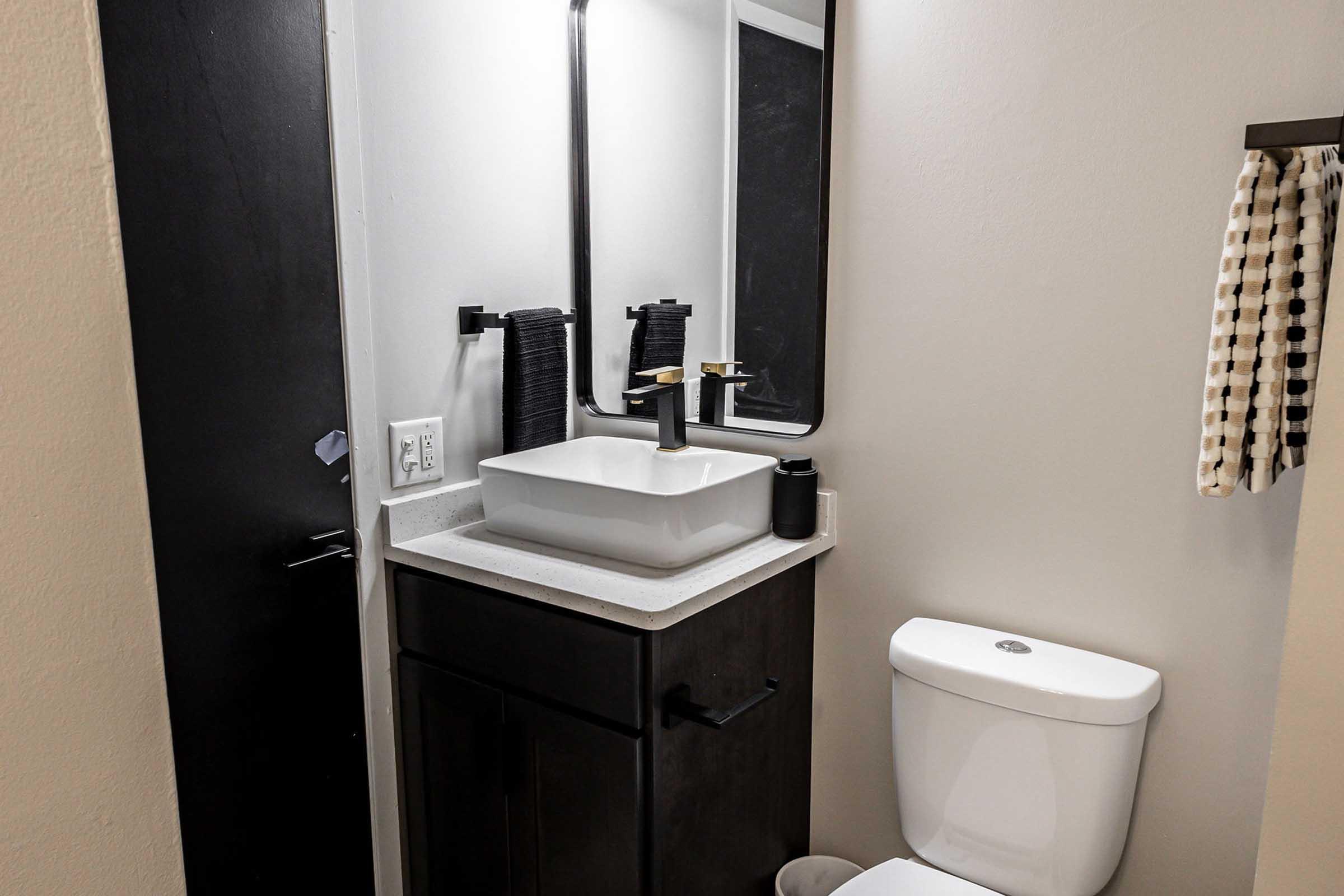 A modern bathroom featuring a sleek black door, a white countertop with a rectangular sink, a black mirror, and decorative towels. The toilet is white, and there is a roll of toilet paper nearby. The overall style is minimalist and contemporary.