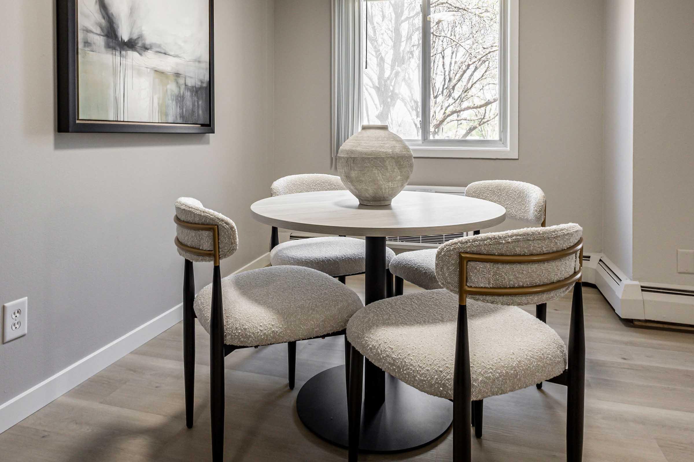 A modern dining area featuring a round white table surrounded by four upholstered chairs with light fabric. A decorative vase sits on the table, and a large abstract painting is hung on the wall. Natural light streams through a nearby window, illuminating the space with a minimalist aesthetic.
