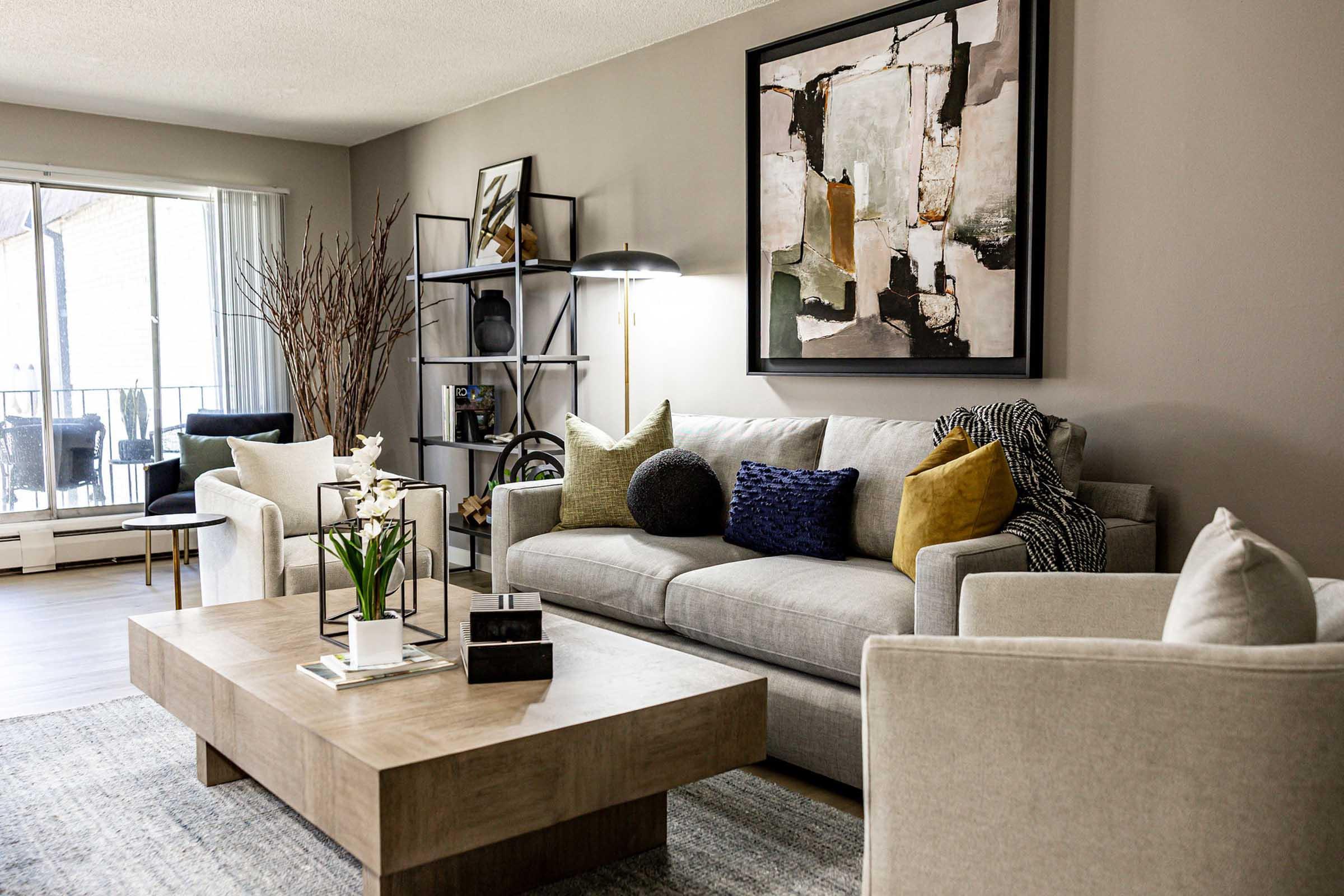 A stylish living room featuring a light gray sofa adorned with colorful pillows, a modern coffee table, and a minimalist shelf displaying decorative items. The walls are painted in a neutral tone, complemented by an abstract artwork above the sofa. Large windows allow natural light to brighten the space, with plants adding a touch of warmth.