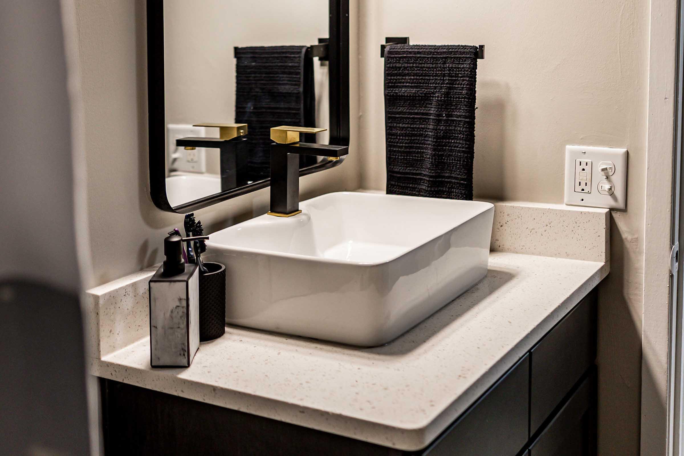 A modern bathroom sink area featuring a stylish white rectangular basin on a light countertop. A black faucet and a black towel hang beside a mirror. There are also decorative items including a sleek black container and toiletries arranged neatly on the countertop.