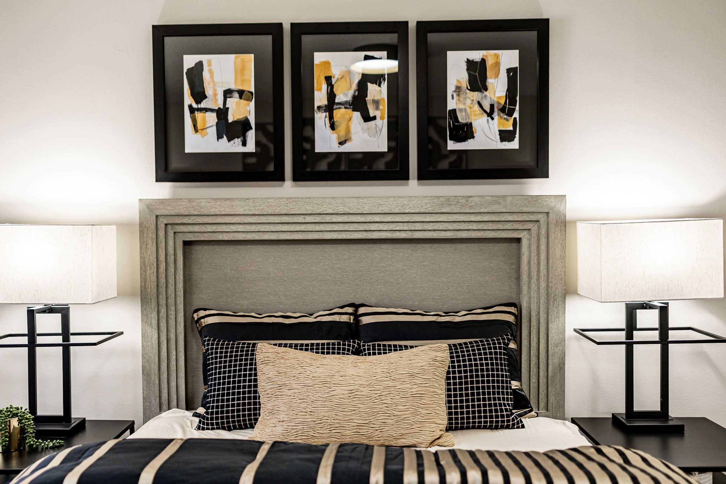 A modern bedroom featuring a stylish headboard with three abstract black and gold artworks framed above it. The bed is adorned with black and white striped pillows and a textured beige pillow. On either side, there are sleek black lamp stands with white lamps, creating a cozy atmosphere.