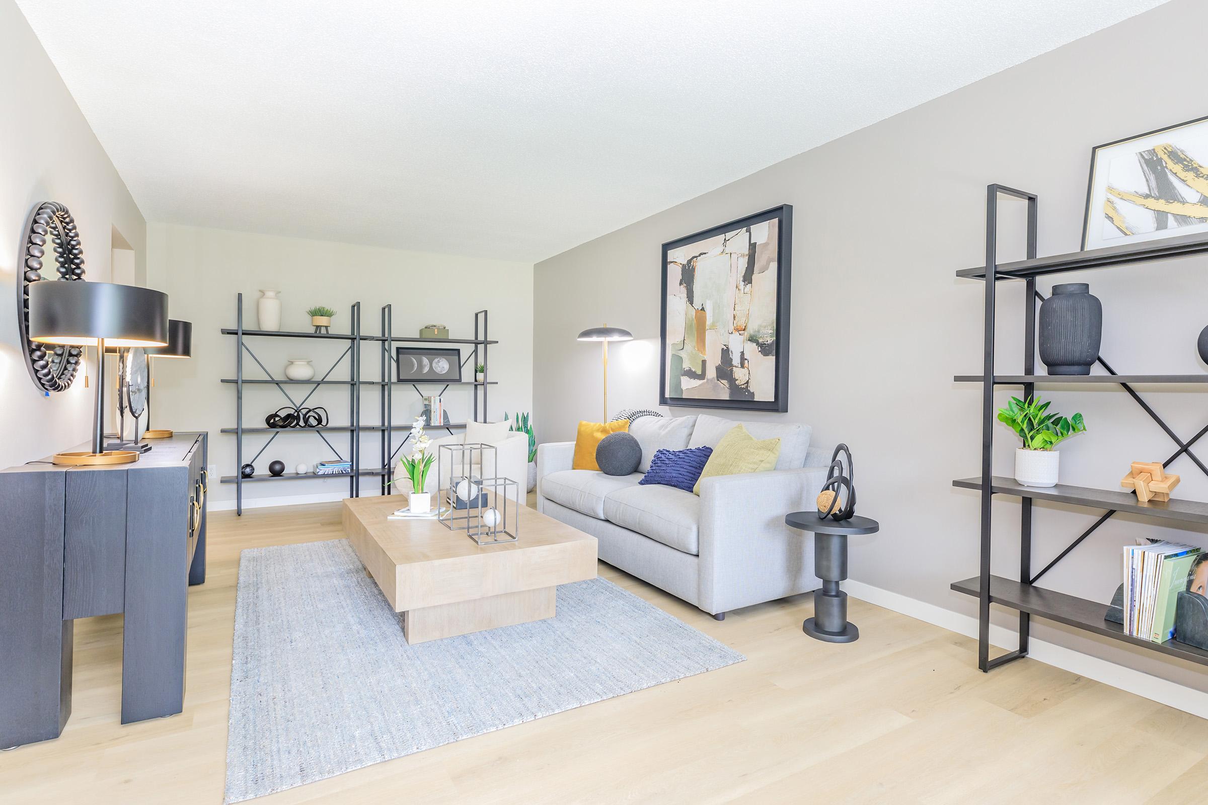 A modern living room featuring a light gray sofa, a wooden coffee table, and decorative shelves with plants and art pieces. The walls are painted a soft gray, and there is a textured area rug on the floor. Lighting includes a floor lamp and table lamp, creating a cozy atmosphere.