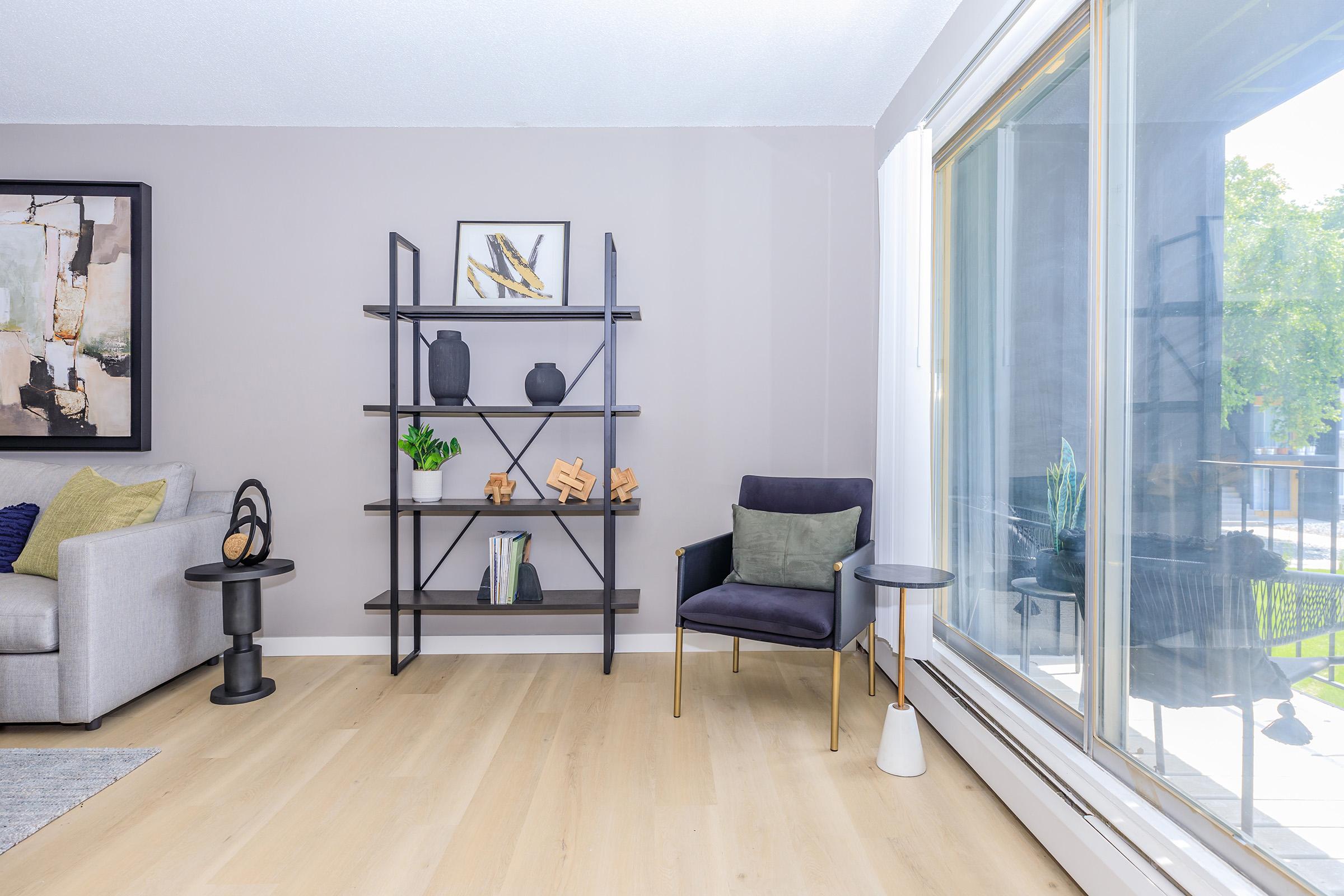 A modern living room featuring a gray sofa with decorative pillows, a black bookshelf with vases and artistic wooden objects, a stylish armchair with cushions, and a small round side table. Large windows let in natural light, illuminating the wooden flooring and creating a bright, inviting atmosphere.