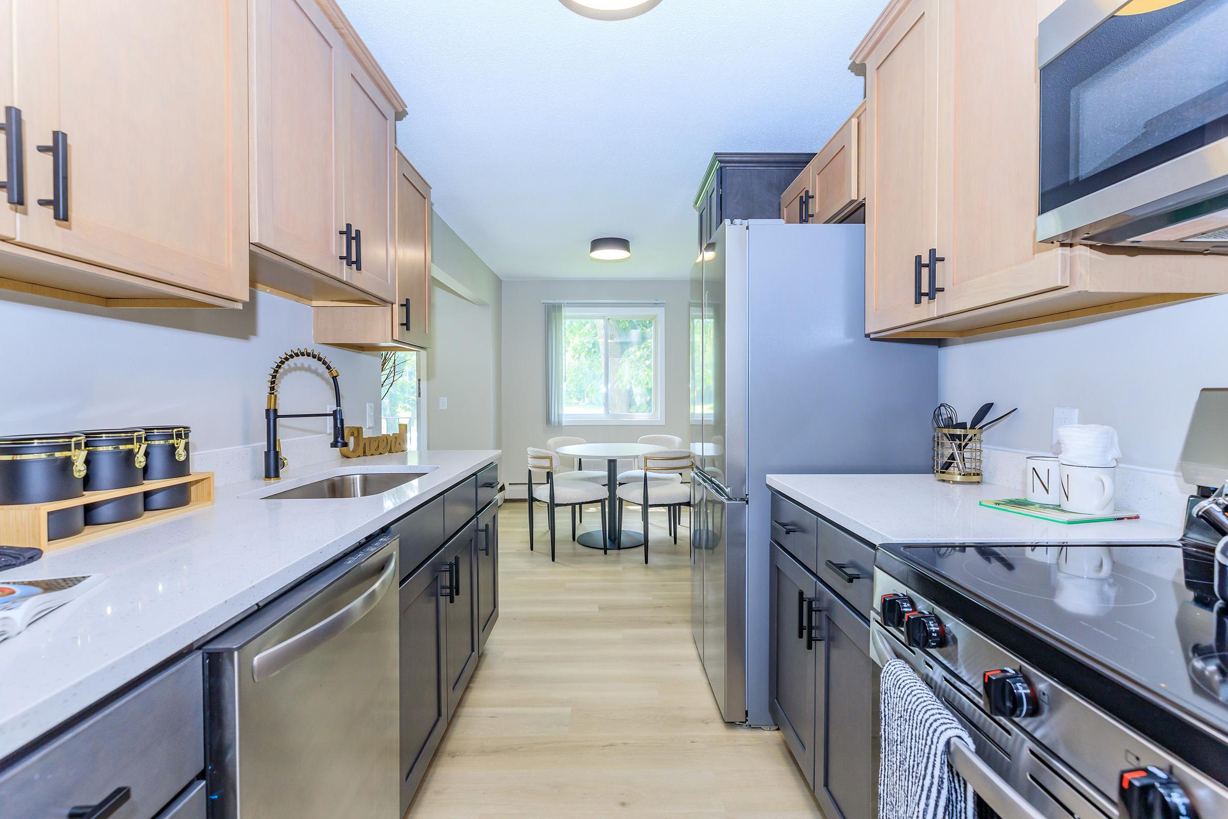 Modern kitchen featuring light wood cabinets, stainless steel appliances, and a sleek gray countertop. The space includes a dining area with a round table and chairs, illuminated by natural light coming through a window. The floors are light wood, creating a bright and inviting atmosphere.