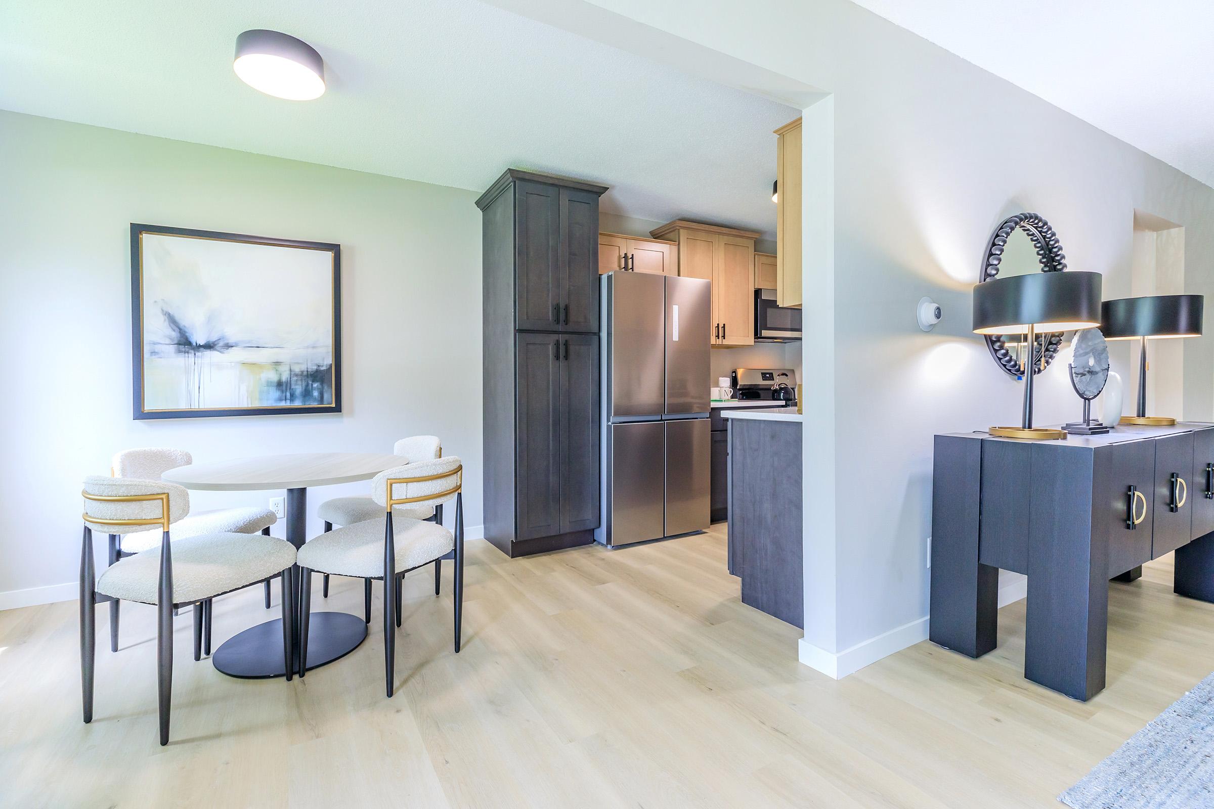 Modern kitchen and dining area featuring a round table with four upholstered chairs, a sleek sideboard, and a stainless steel refrigerator. The walls are painted in a light color, complemented by an abstract painting and warm, natural light coming in from the windows.