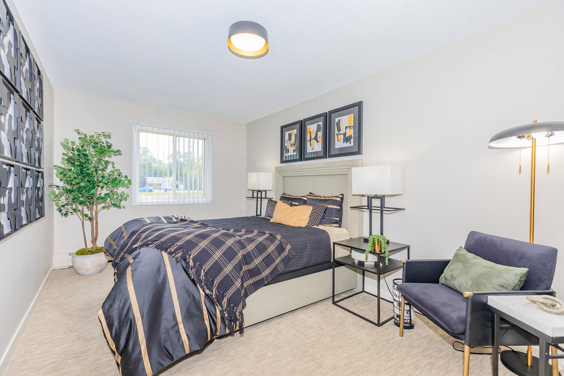 A cozy bedroom featuring a neatly made bed with dark bedding, two bedside lamps, and framed artwork on the walls. A small tree adds greenery, and a chair with a cushion complements the decor. Natural light enters through a window with vertical blinds. The room has a modern and inviting atmosphere.