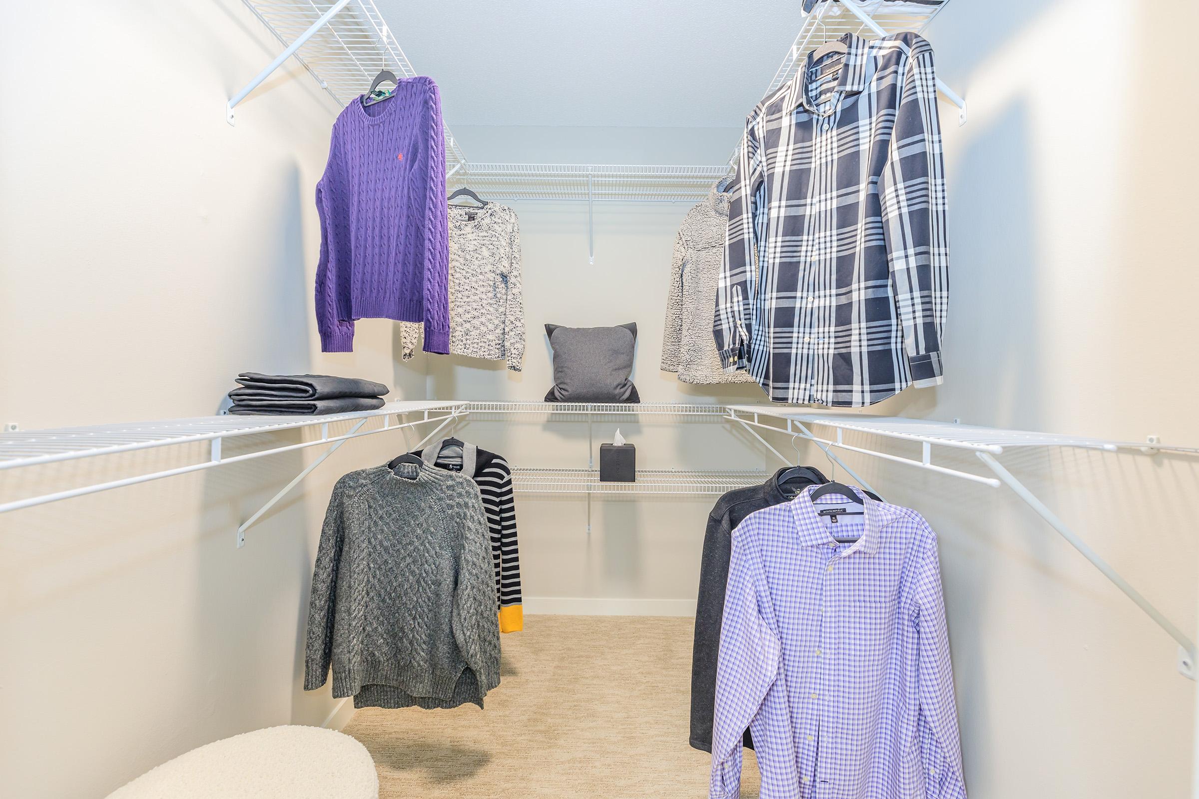 A neatly organized walk-in closet featuring various hanging clothes, including sweaters and shirts in different colors and patterns. Two shelves are filled with folded items and a cushion, against a light-colored wall and soft carpet flooring.