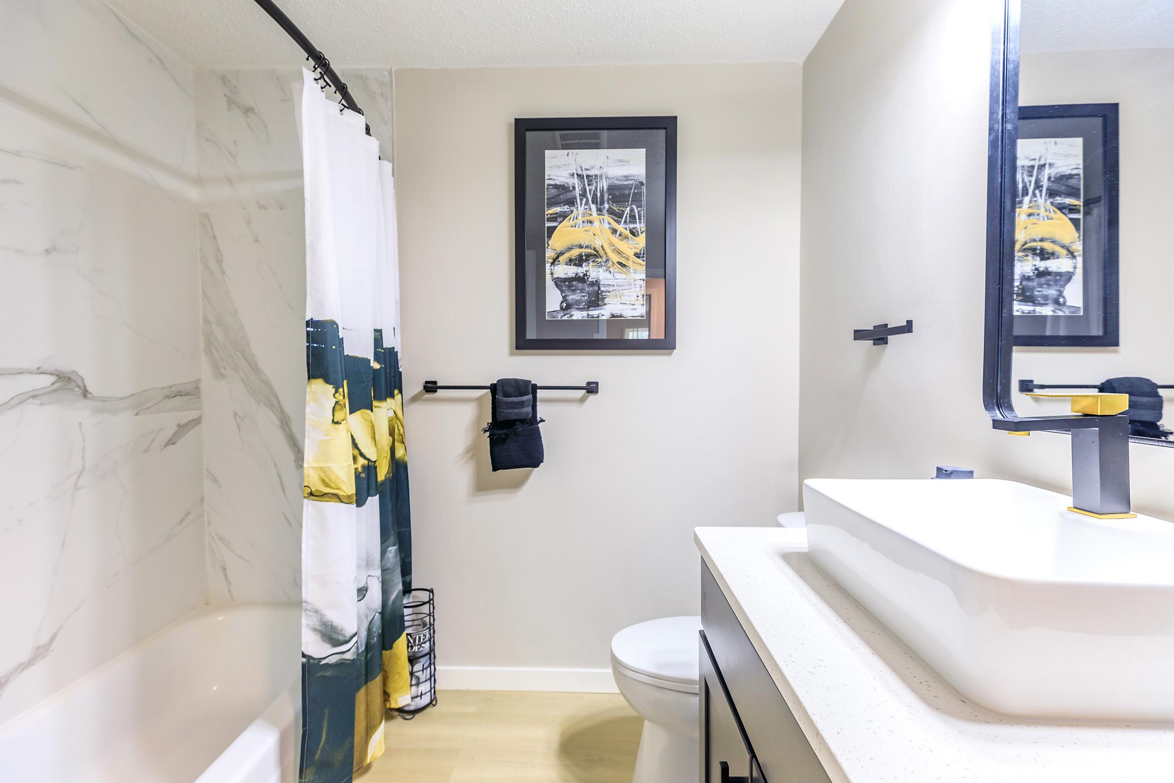 A modern bathroom featuring a white bathtub with a shower curtain displaying a colorful design. There is a minimalist black and white wall art piece above a sleek sink with a stylish faucet. A toilet is visible, and a towel is hanging on a bar beside a small wastebasket. The walls have a marble effect.
